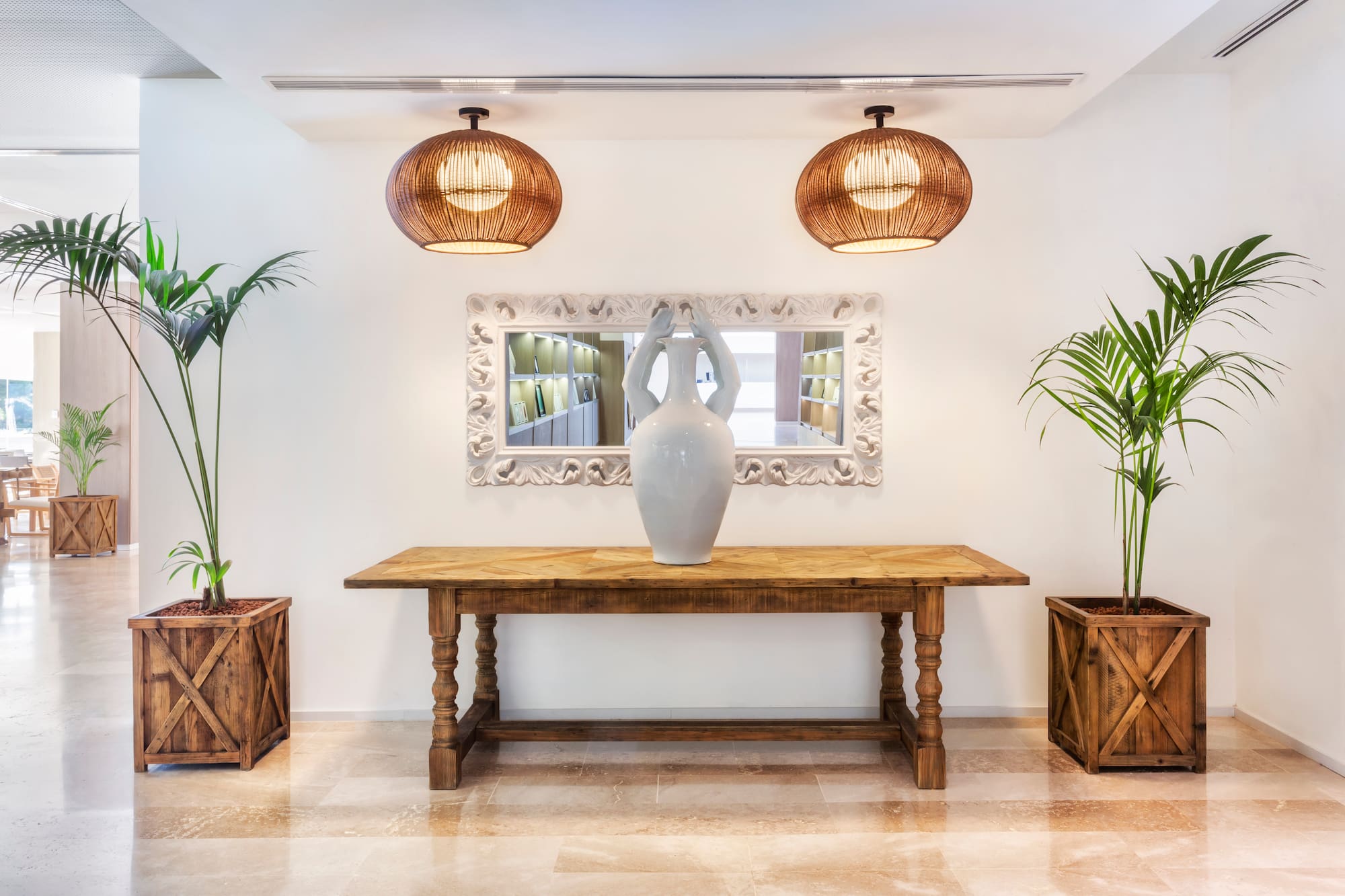 Lobby decor: wooden console table, ornate mirror, white vase, and palm plants.