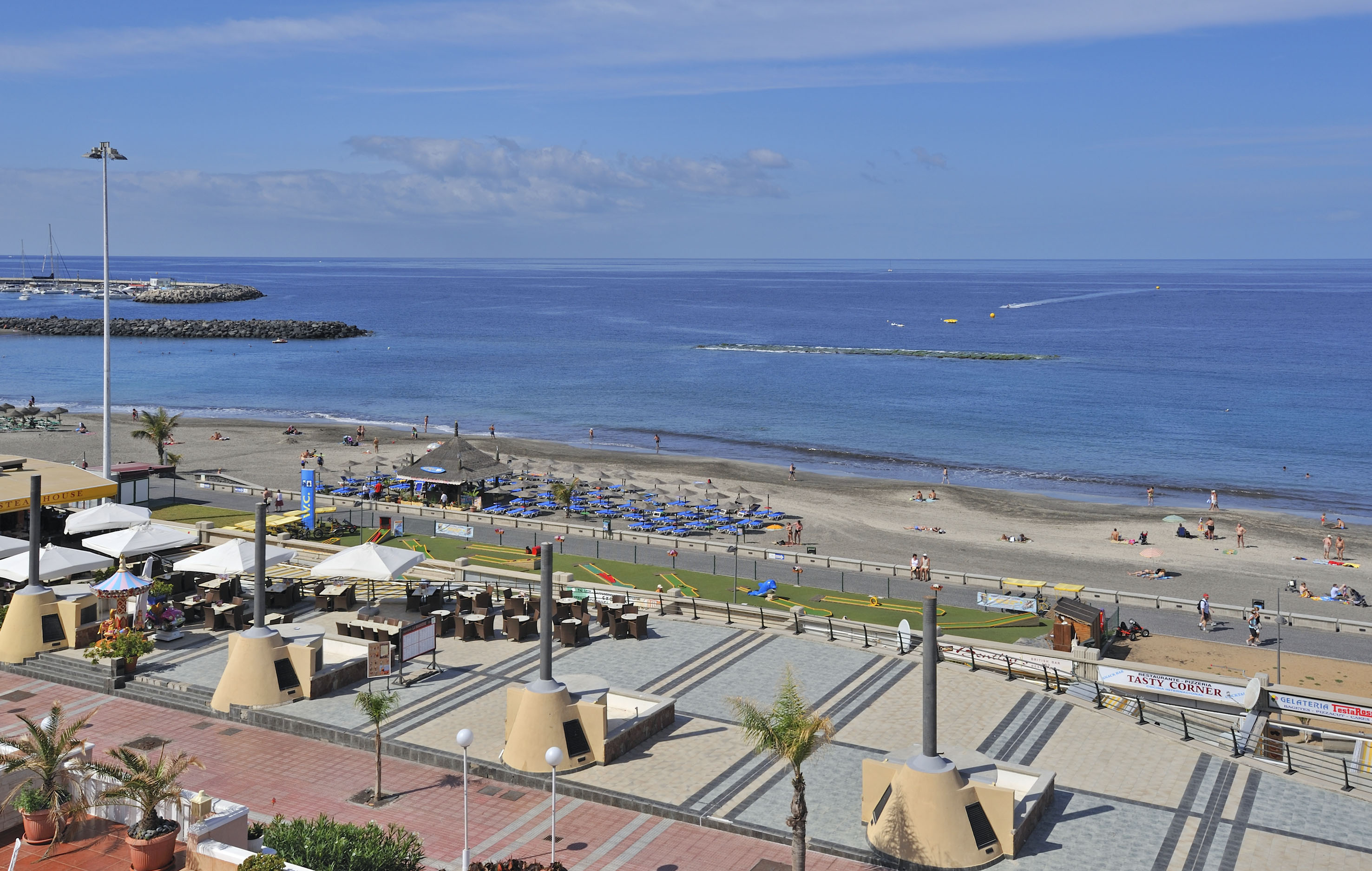 Sunny beach resort with busy promenade, blue ocean, and sunbathers.