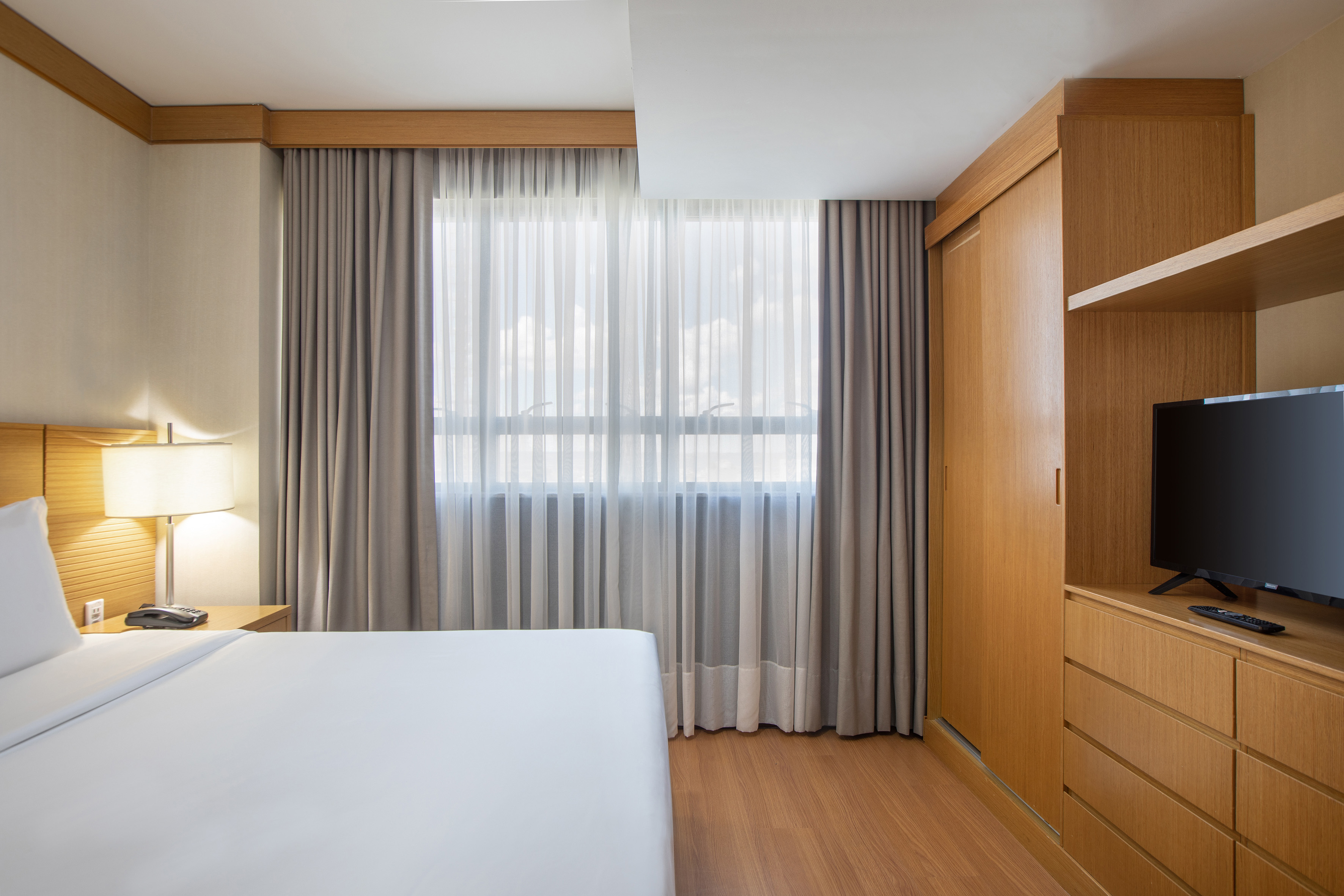 Bright modern hotel bedroom with white bed, wooden wardrobe, window, and TV.