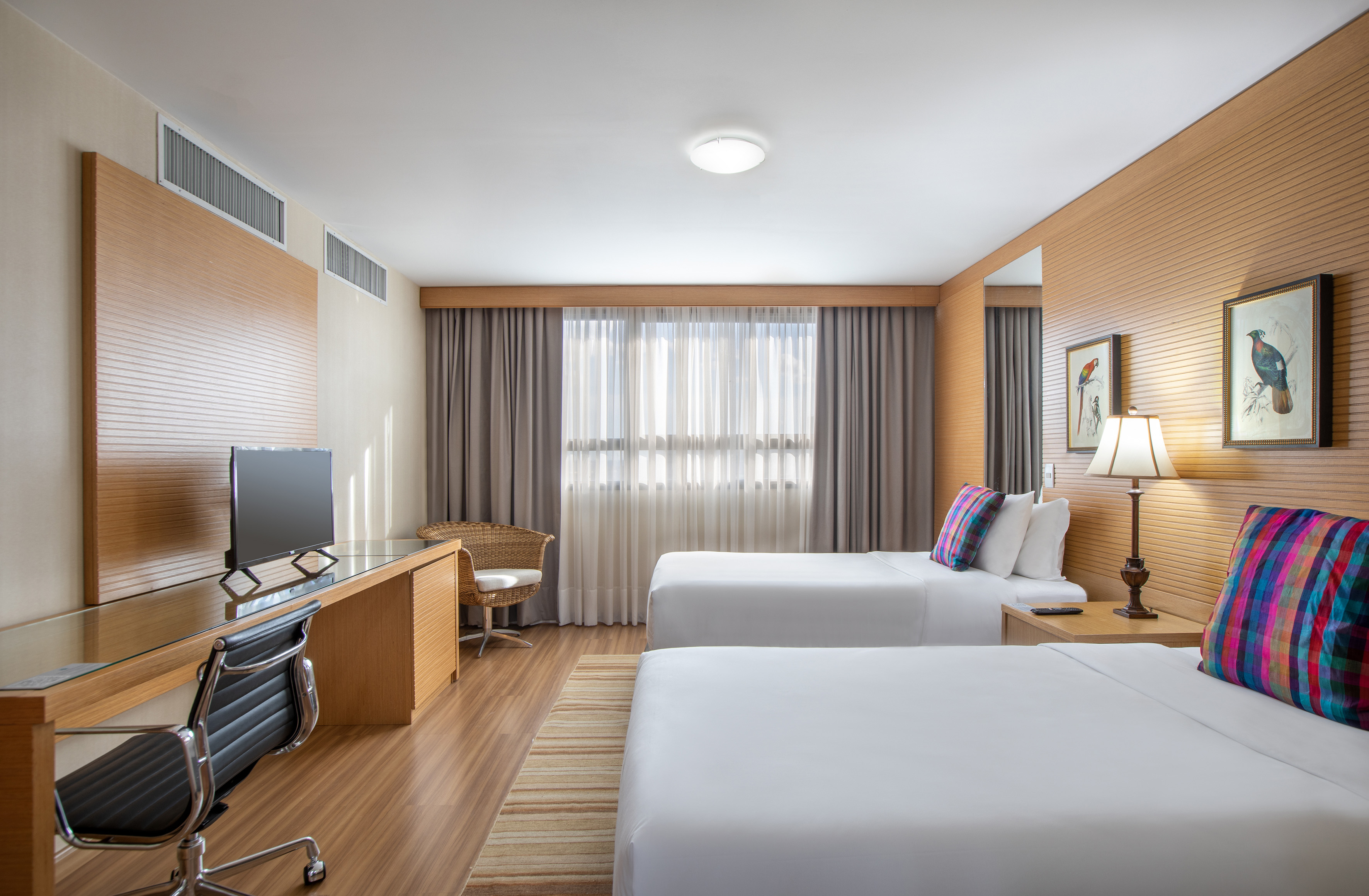 Modern twin hotel room with two beds, wooden desk, TV, and patterned cushions.