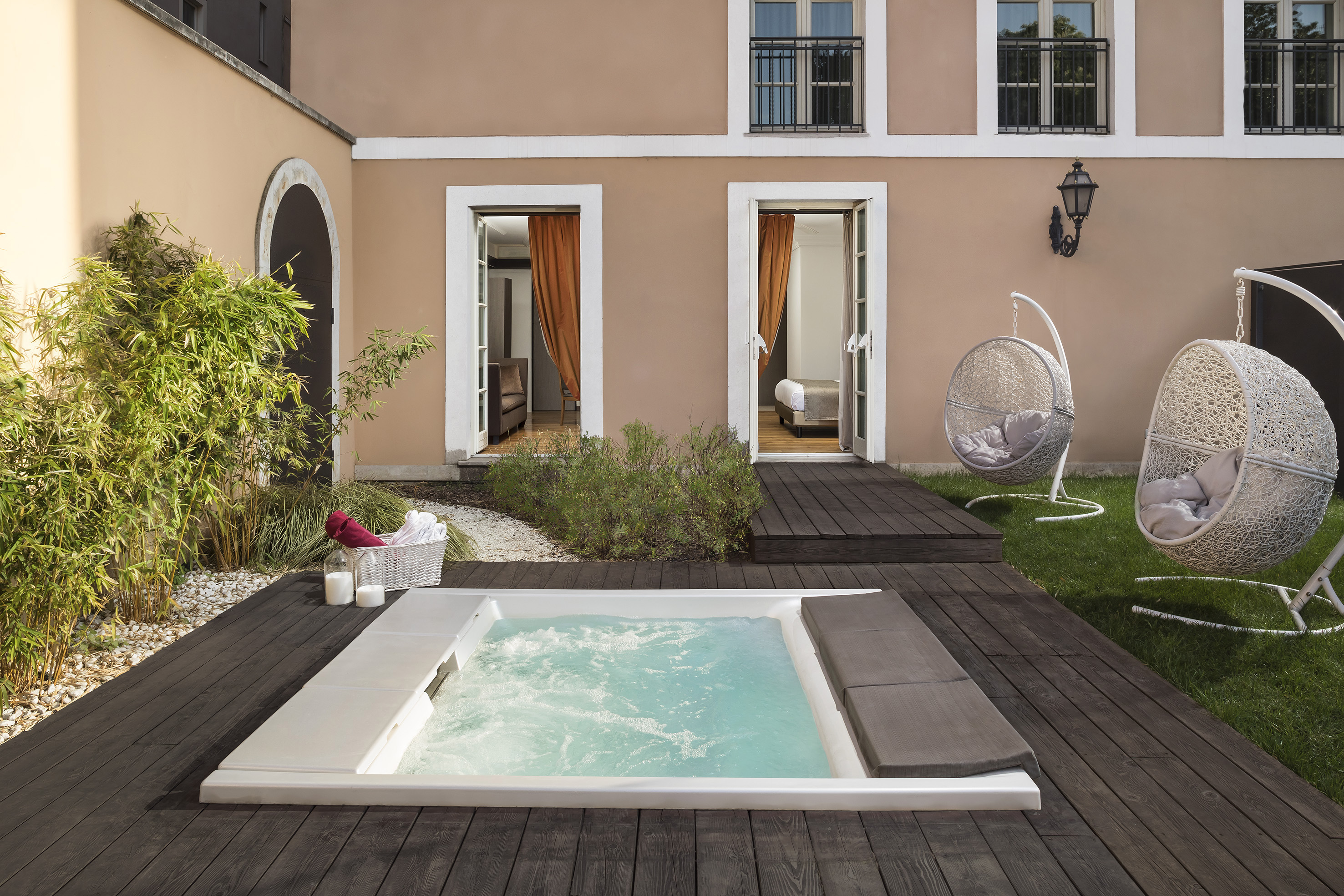Sunny hotel patio with hot tub, wooden deck, two hanging chairs, and open rooms.