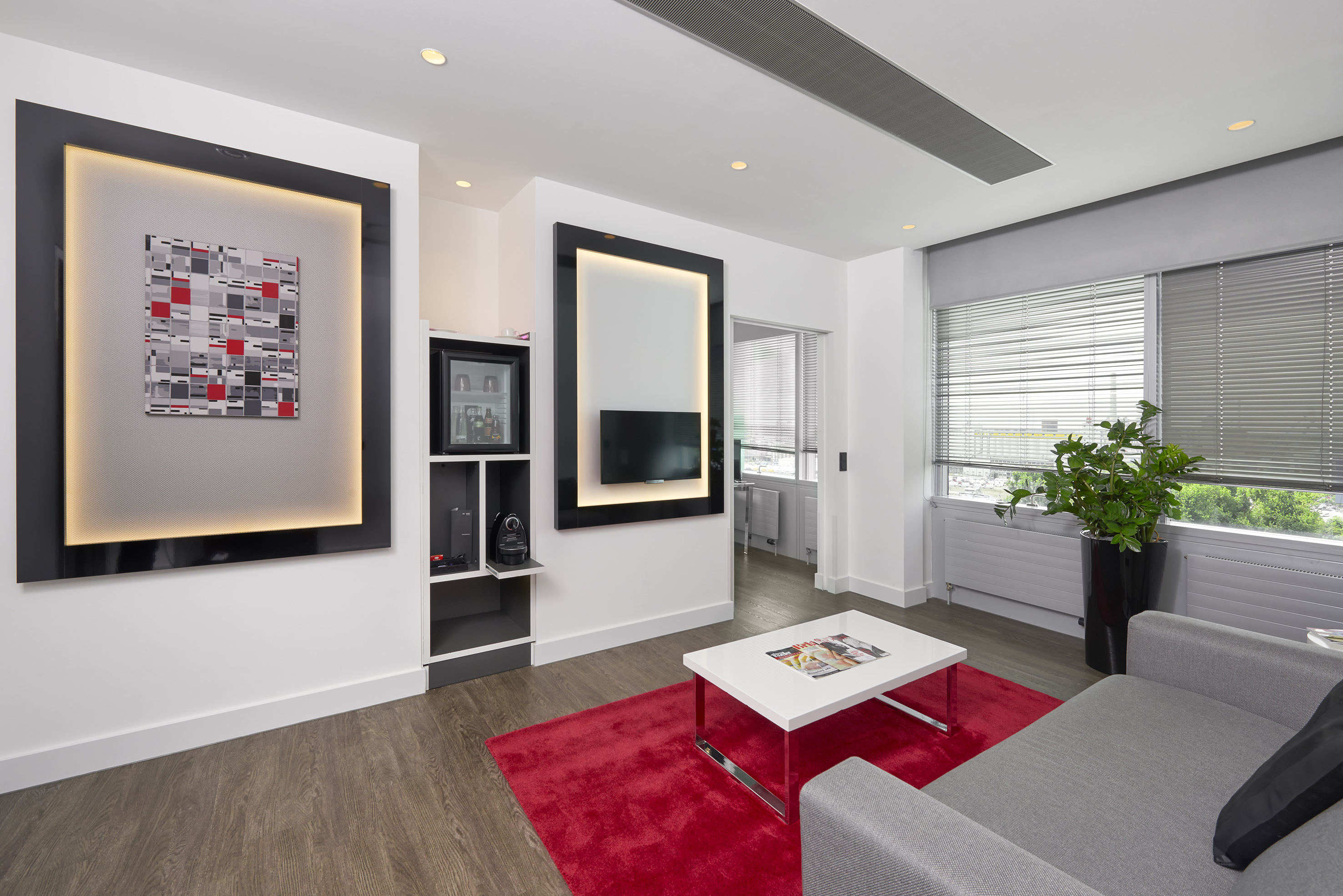Modern hotel living room, gray sofa, red rug, abstract art, and wall TV.
