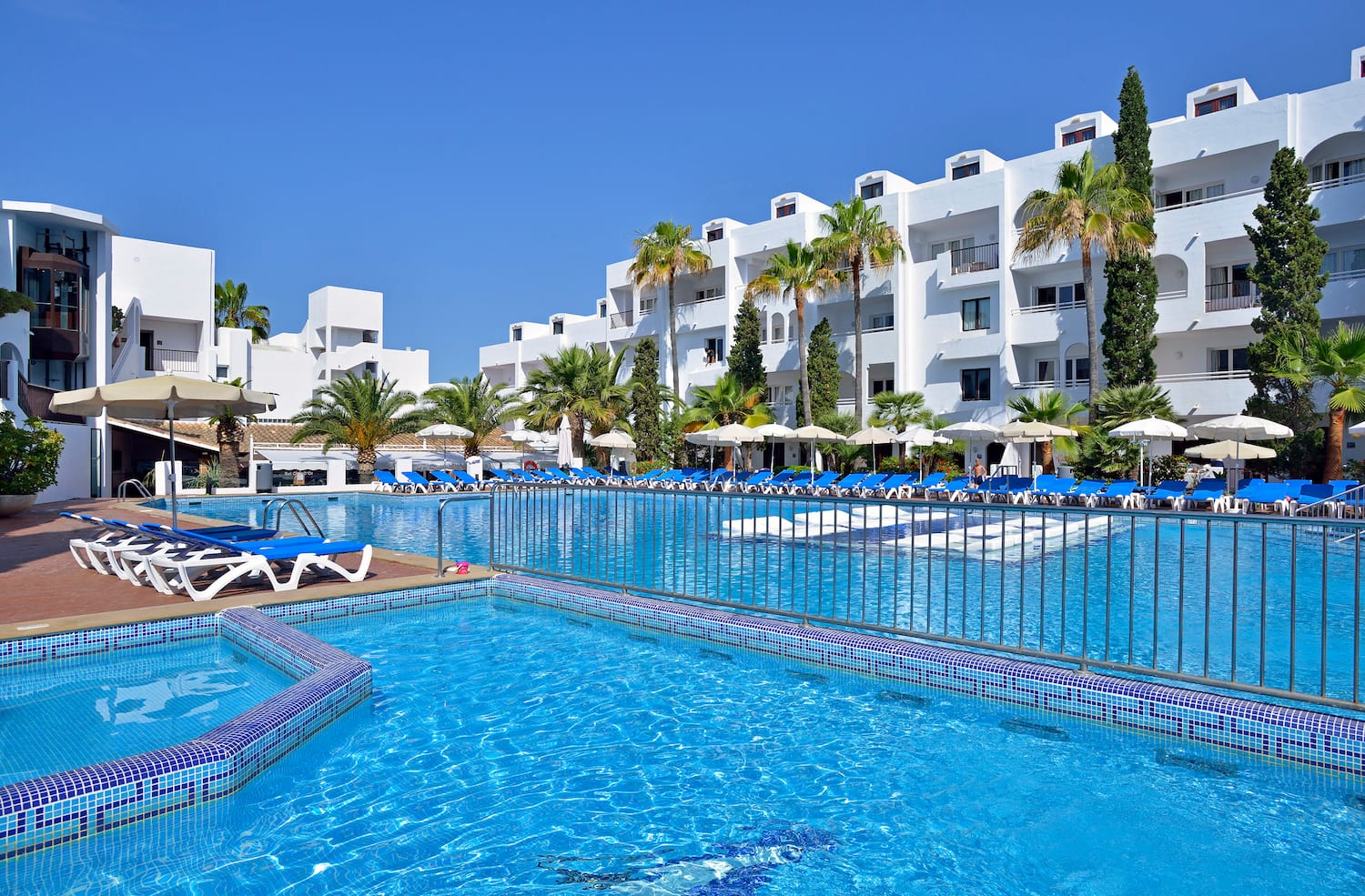 a pool with lounge chairs and a fence in front of a white building