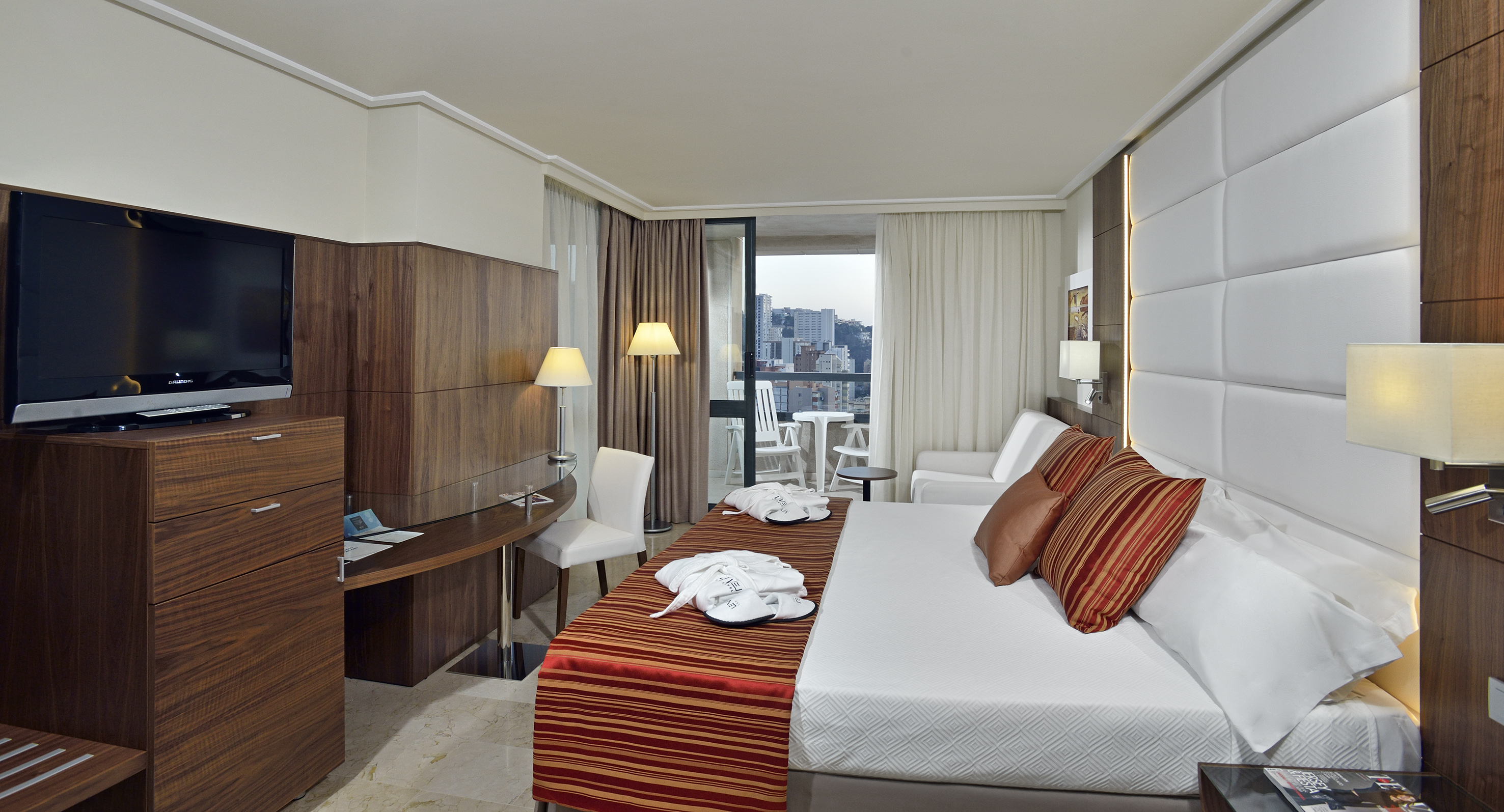 Modern hotel room with bed, desk, TV, and balcony overlooking city skyline.