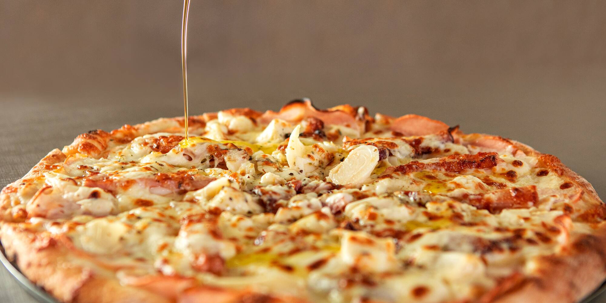 Pizza with melted cheese, meat toppings, olive oil drizzling from green bottle.