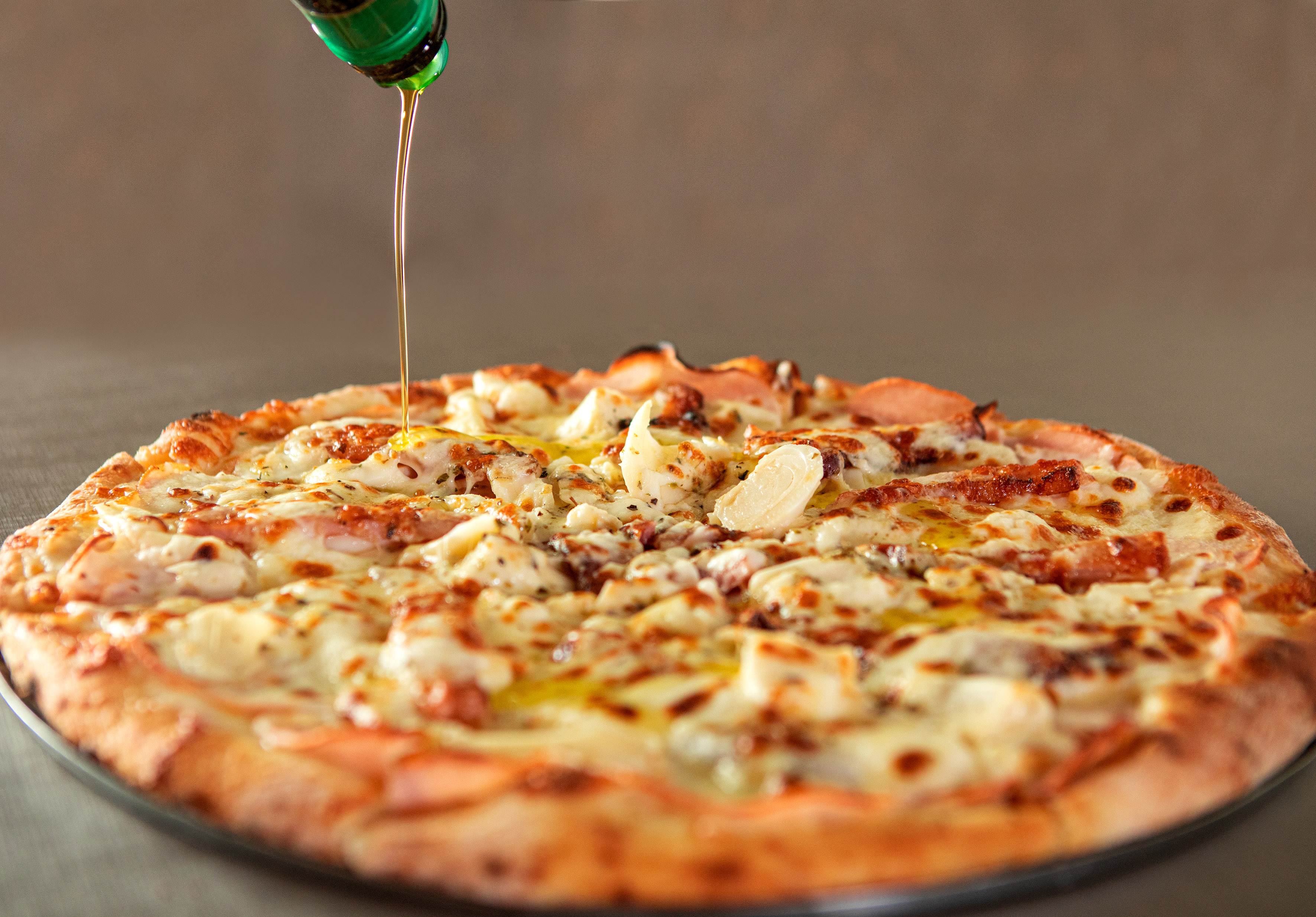Pizza with melted cheese, meat toppings, olive oil drizzling from green bottle.