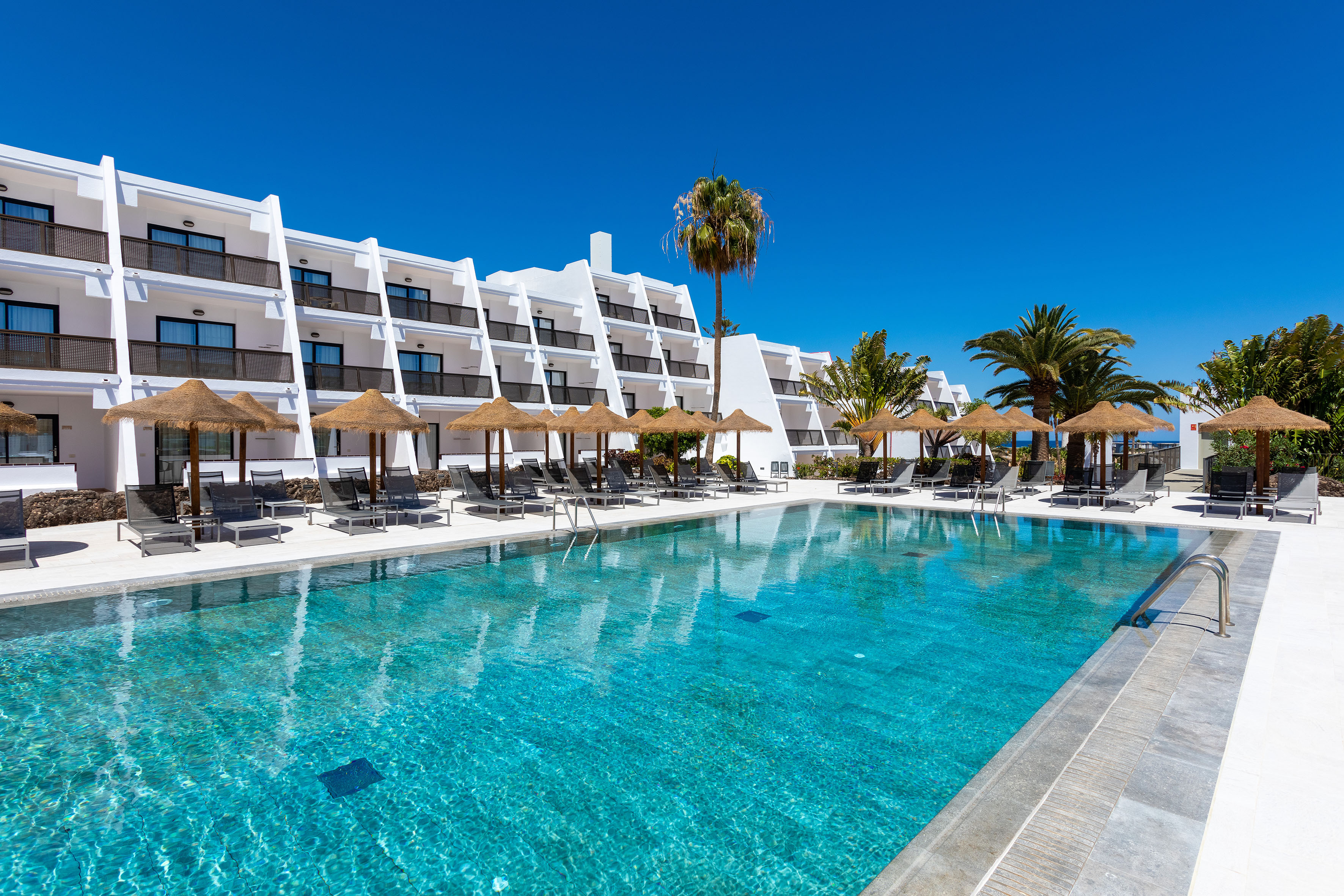 Resort hotel pool with white building, palm trees, sun loungers, and umbrellas.
