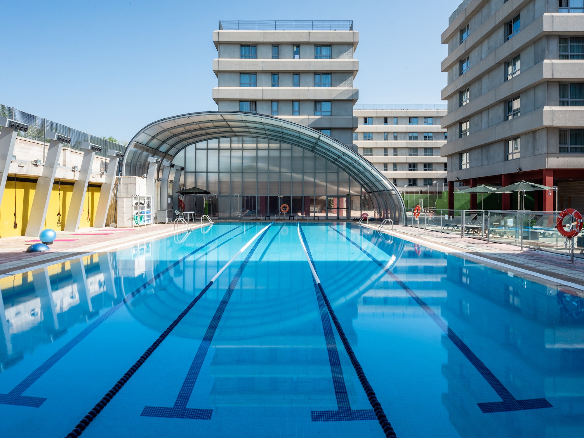 Piscina exterior azul con carriles, estructura acristalada y edificios.