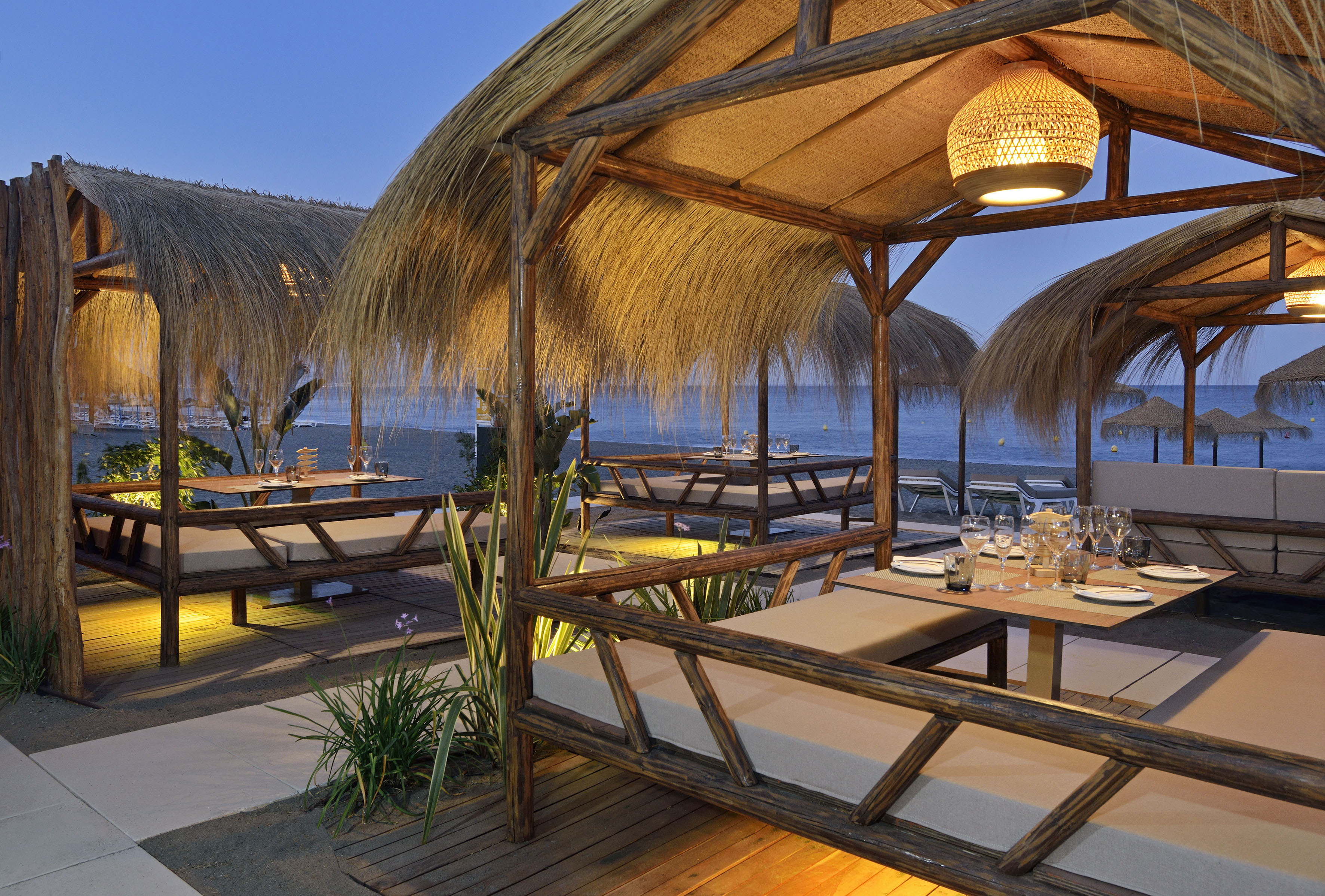 Illuminated tropical beachfront restaurant with thatched huts and sea view.