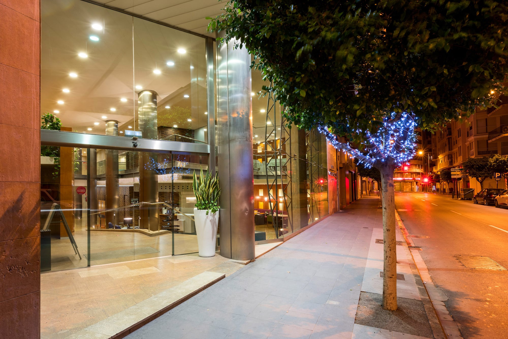 Modern glass hotel entrance at night, illuminated tree, city sidewalk.