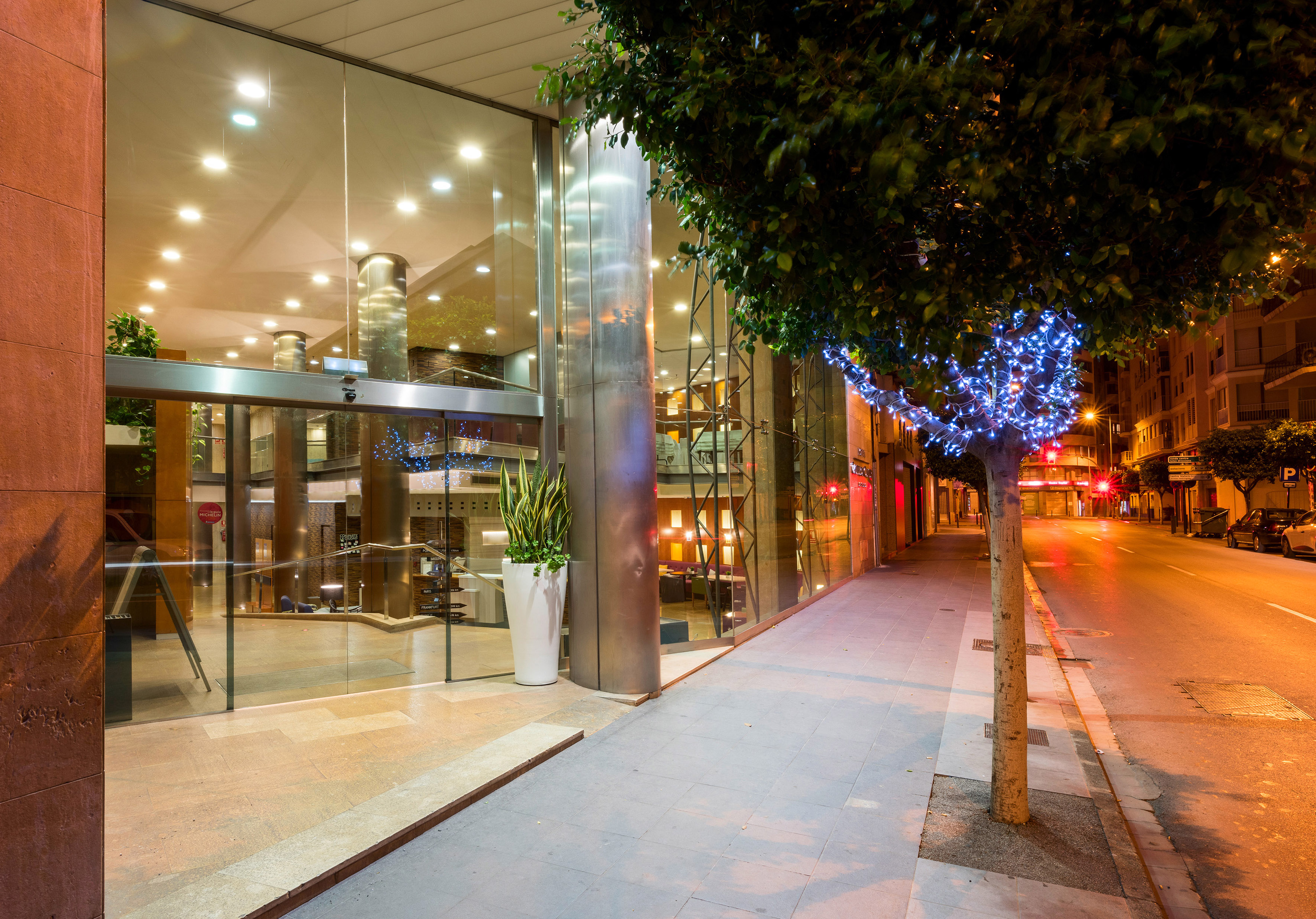 Modern glass hotel entrance at night, illuminated tree, city sidewalk.