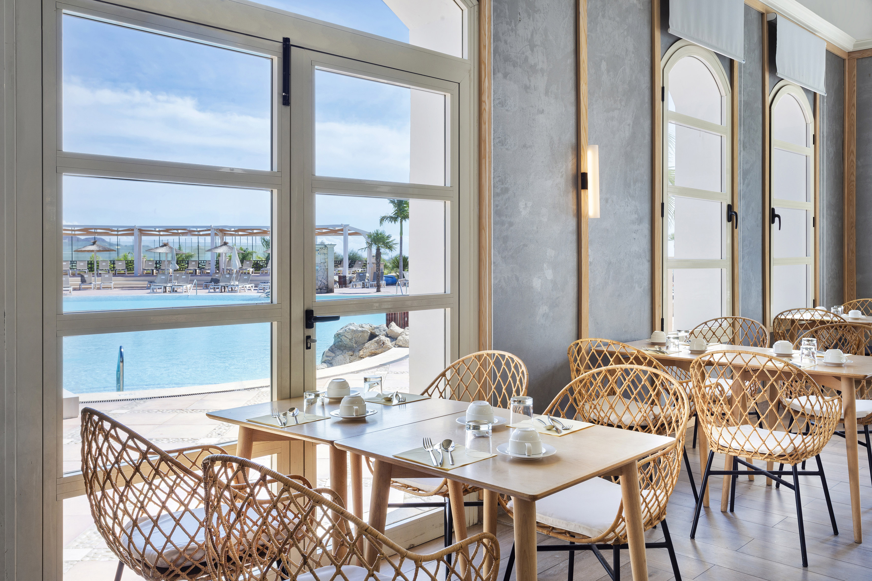 Hotel dining room with pool view, light wood tables, and woven rattan chairs.