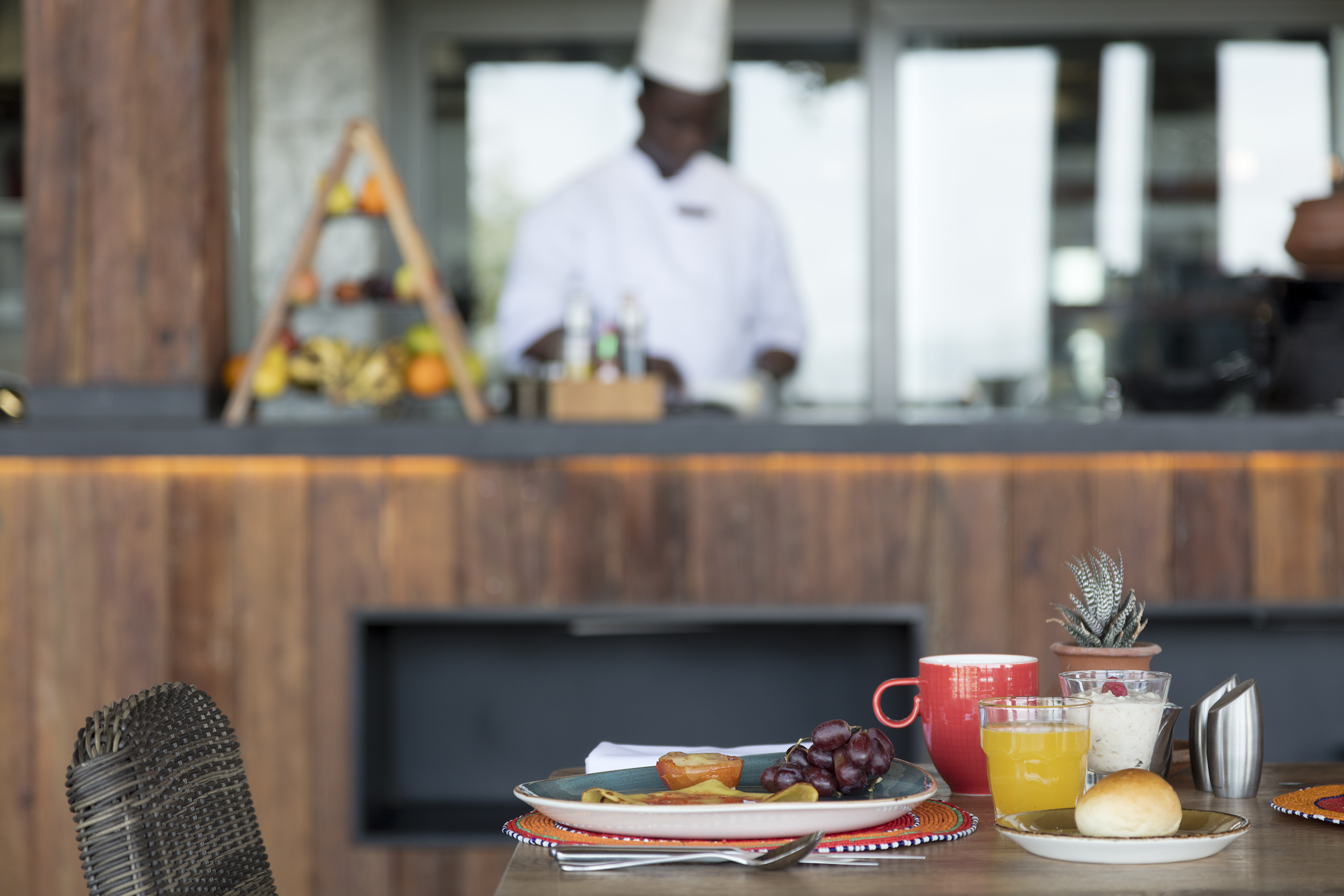 Hotel breakfast table with fresh fruit, juice, pastry; chef working in background.