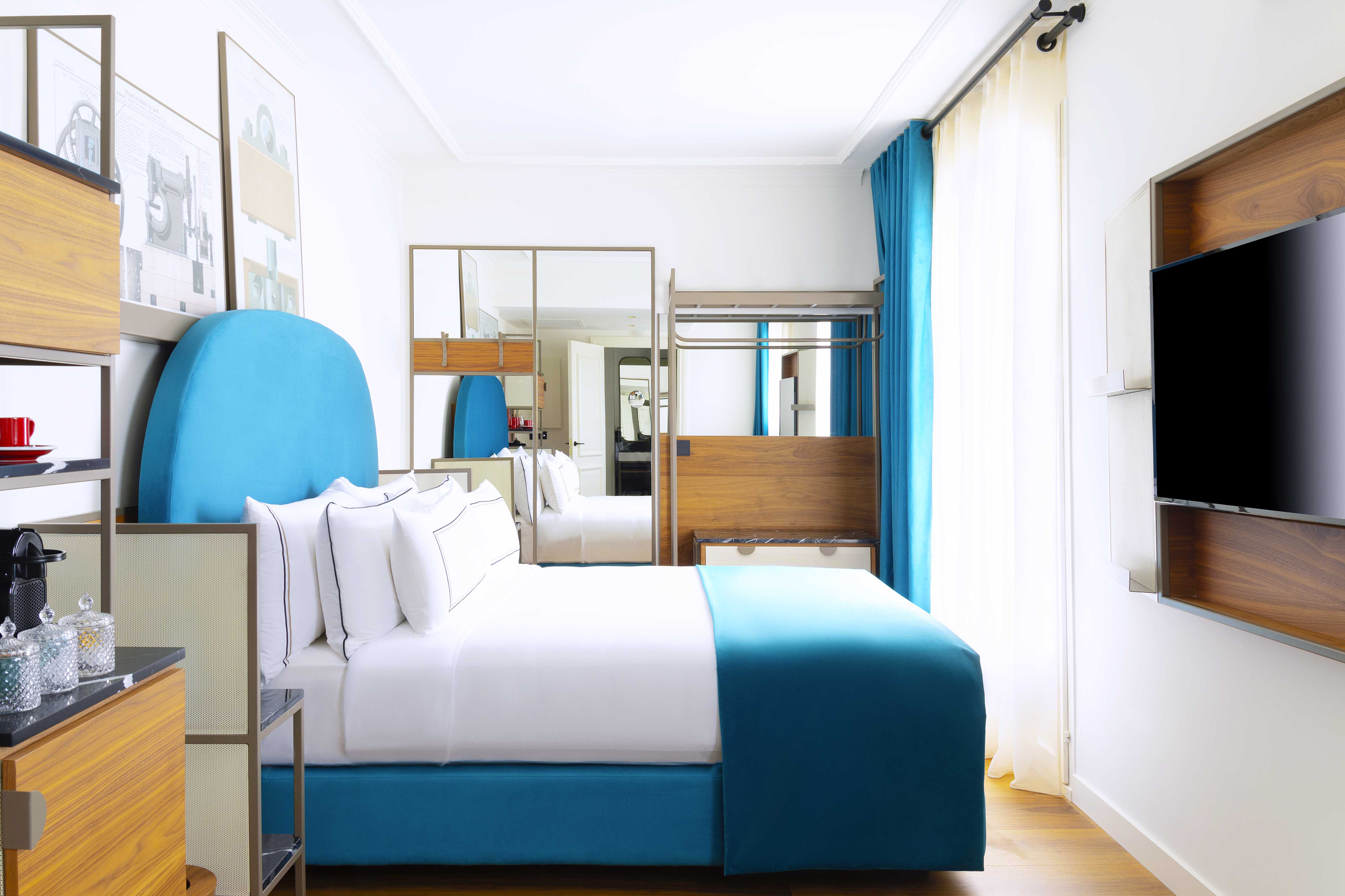 Bright modern hotel room with blue bed, white linen, wood furniture, TV.
