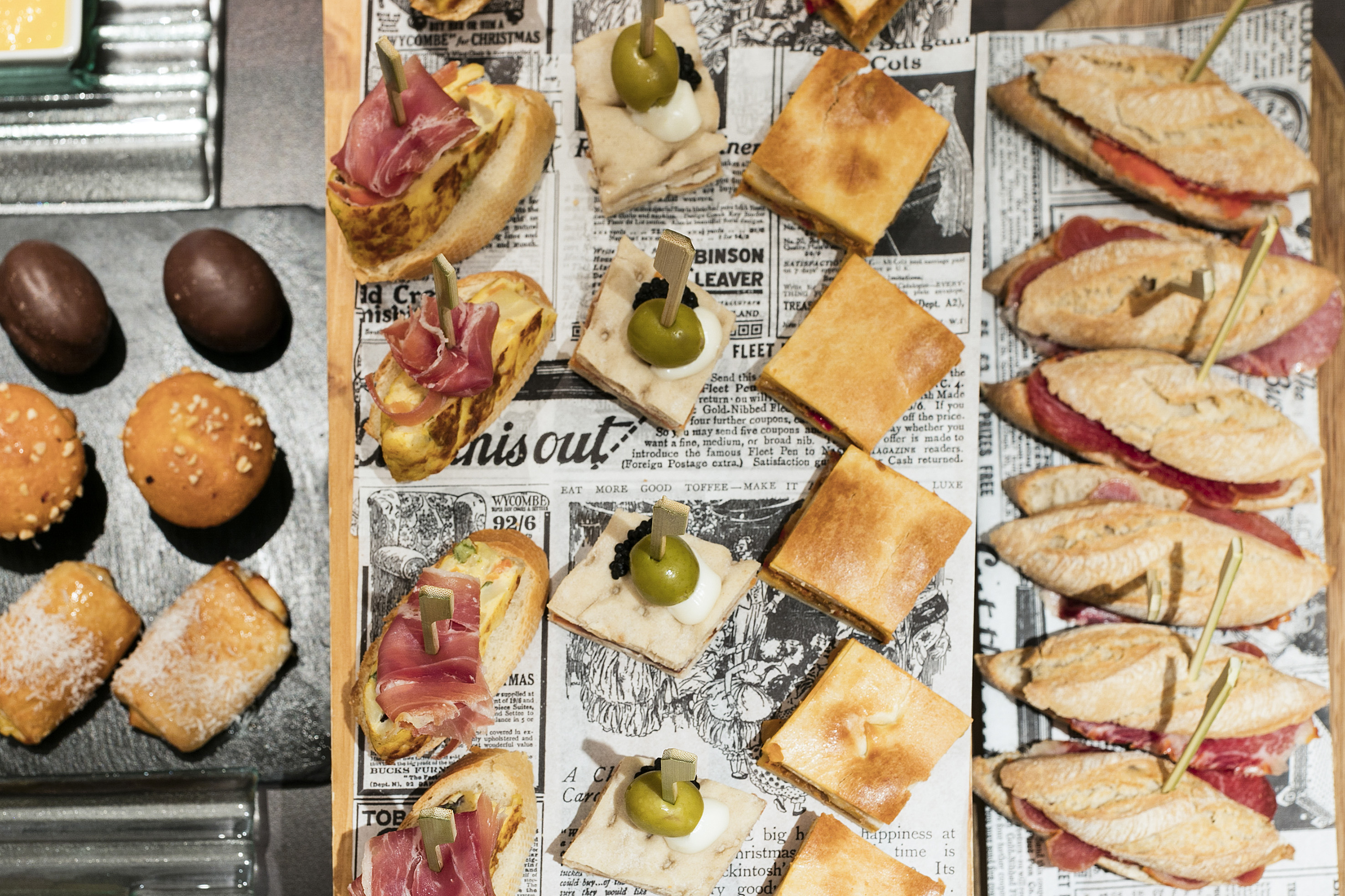 Spanish tapas, mini sandwiches, and pastries served on vintage newspaper.