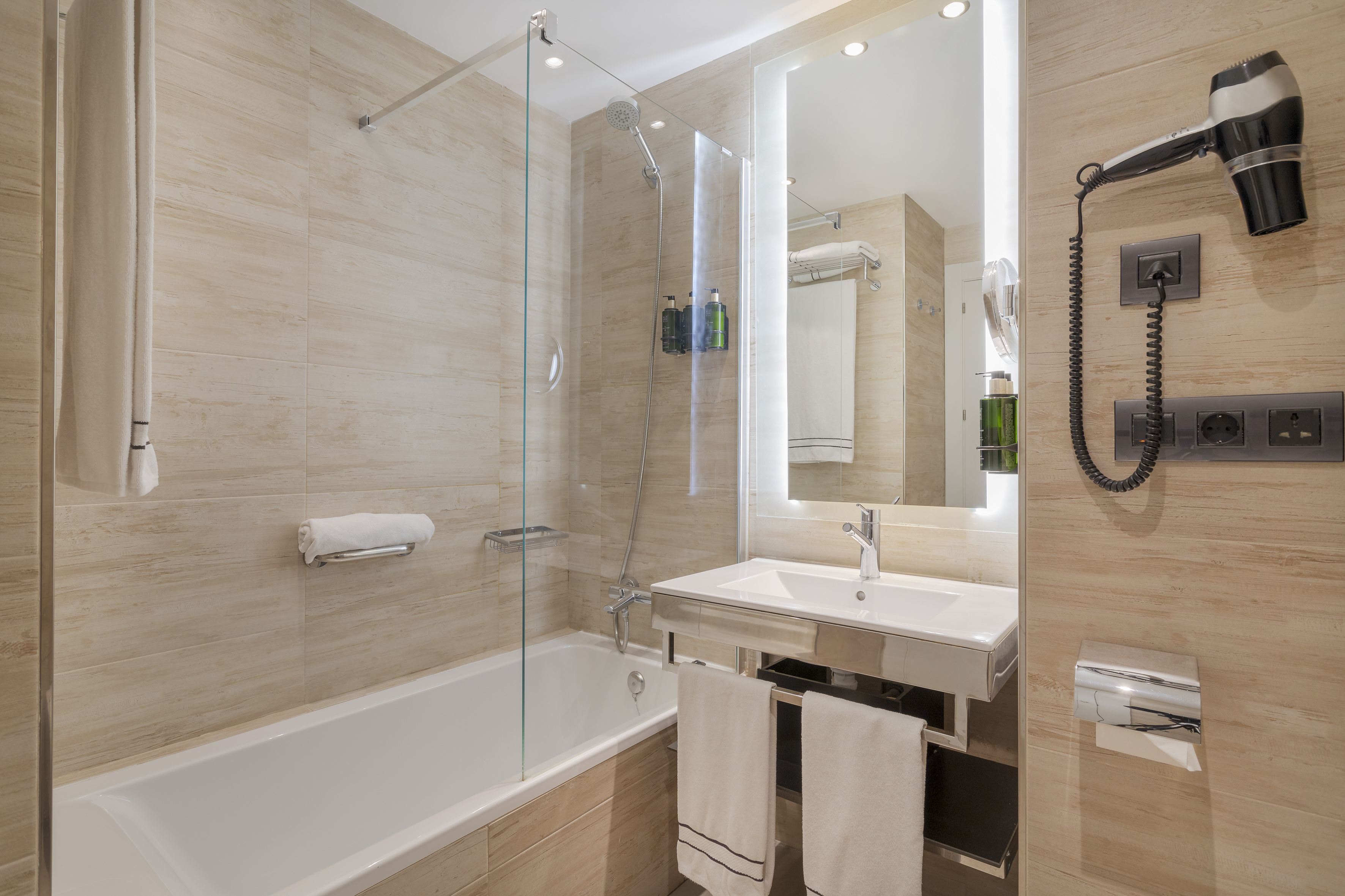 Modern hotel bathroom with bathtub, shower, vanity, and mounted hairdryer.
