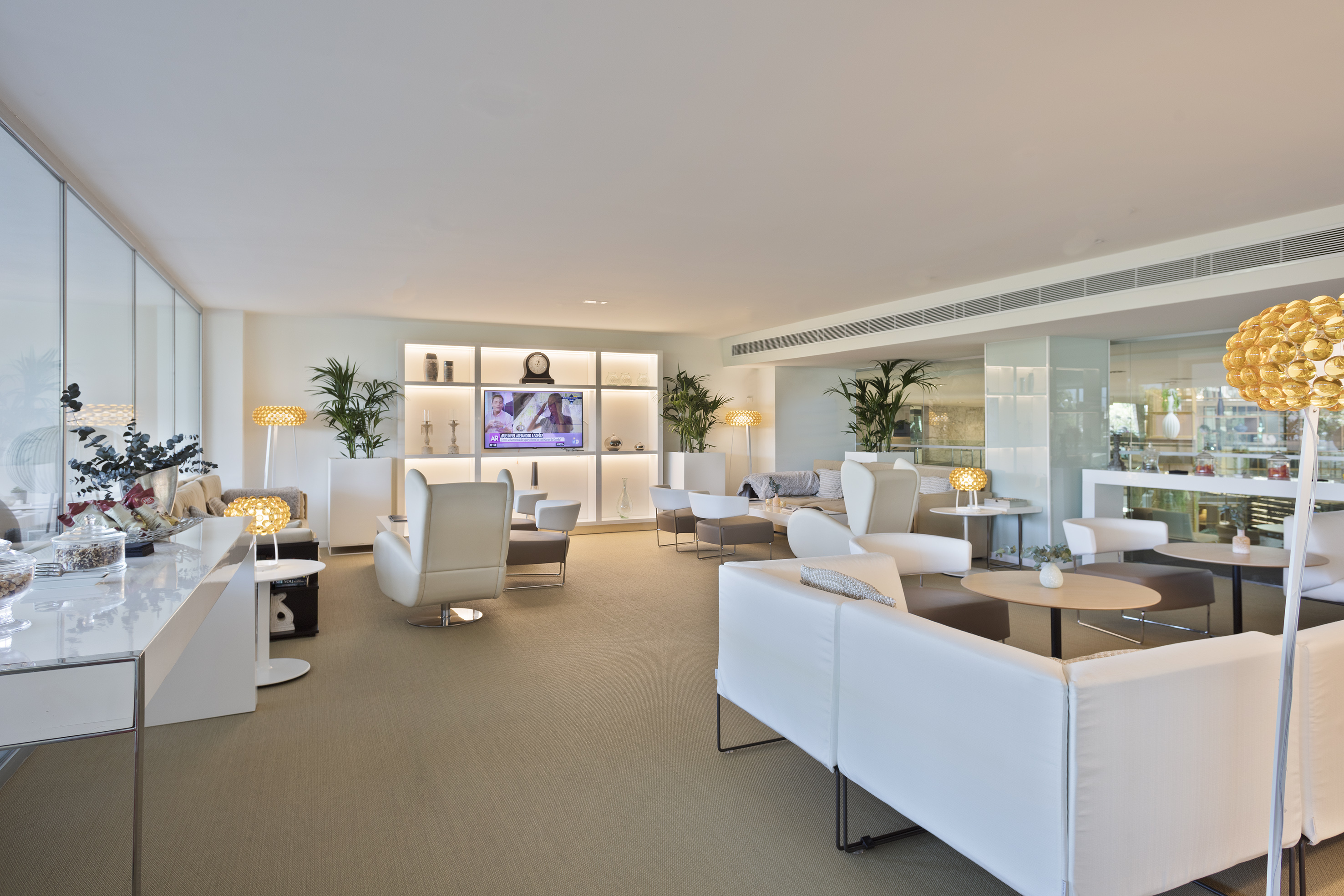 Modern hotel lounge with white furniture, TV, and decorative plants.
