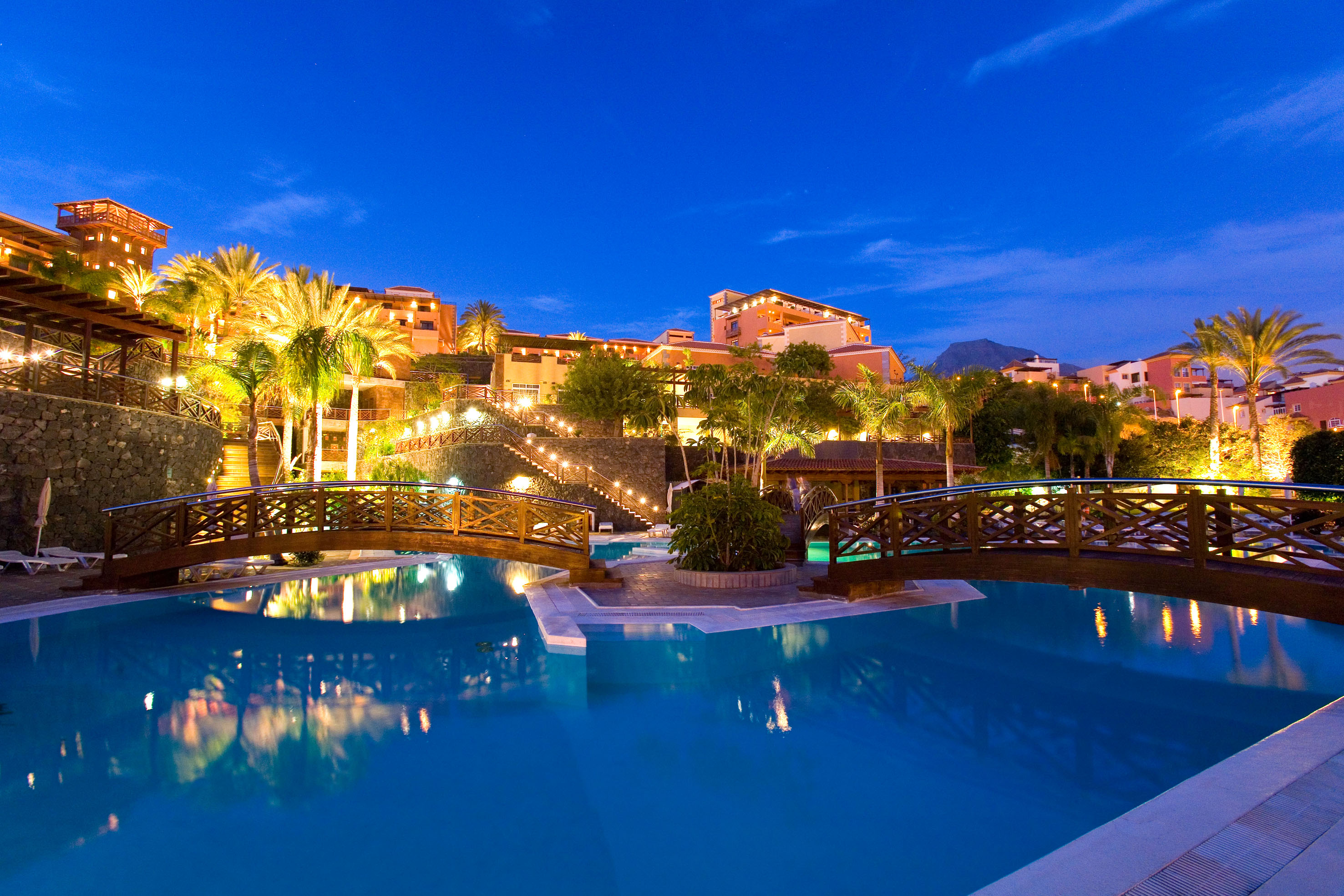 Tropical resort hotel with illuminated pool, palm trees, and bridges at dusk.