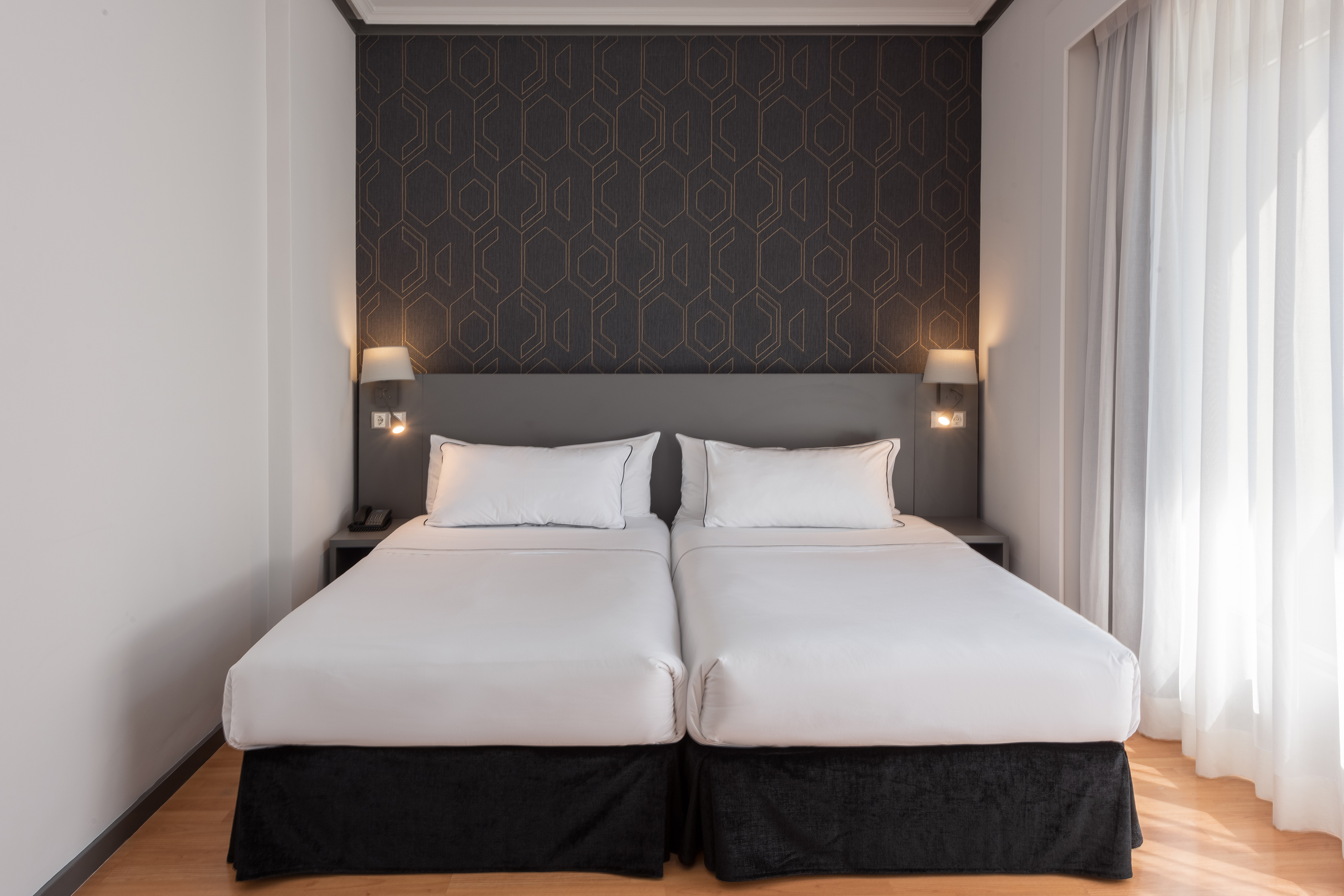 Hotel room with two twin beds, geometric wallpaper, headboard, and sheer curtains.