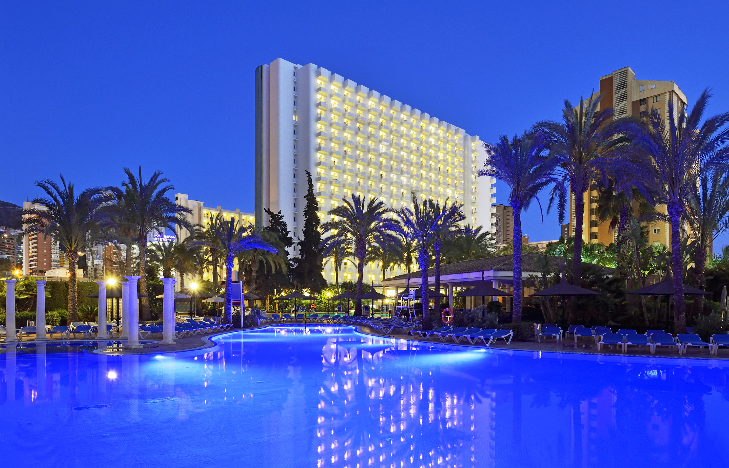 Illuminated resort hotel with blue swimming pool and palm trees at night.