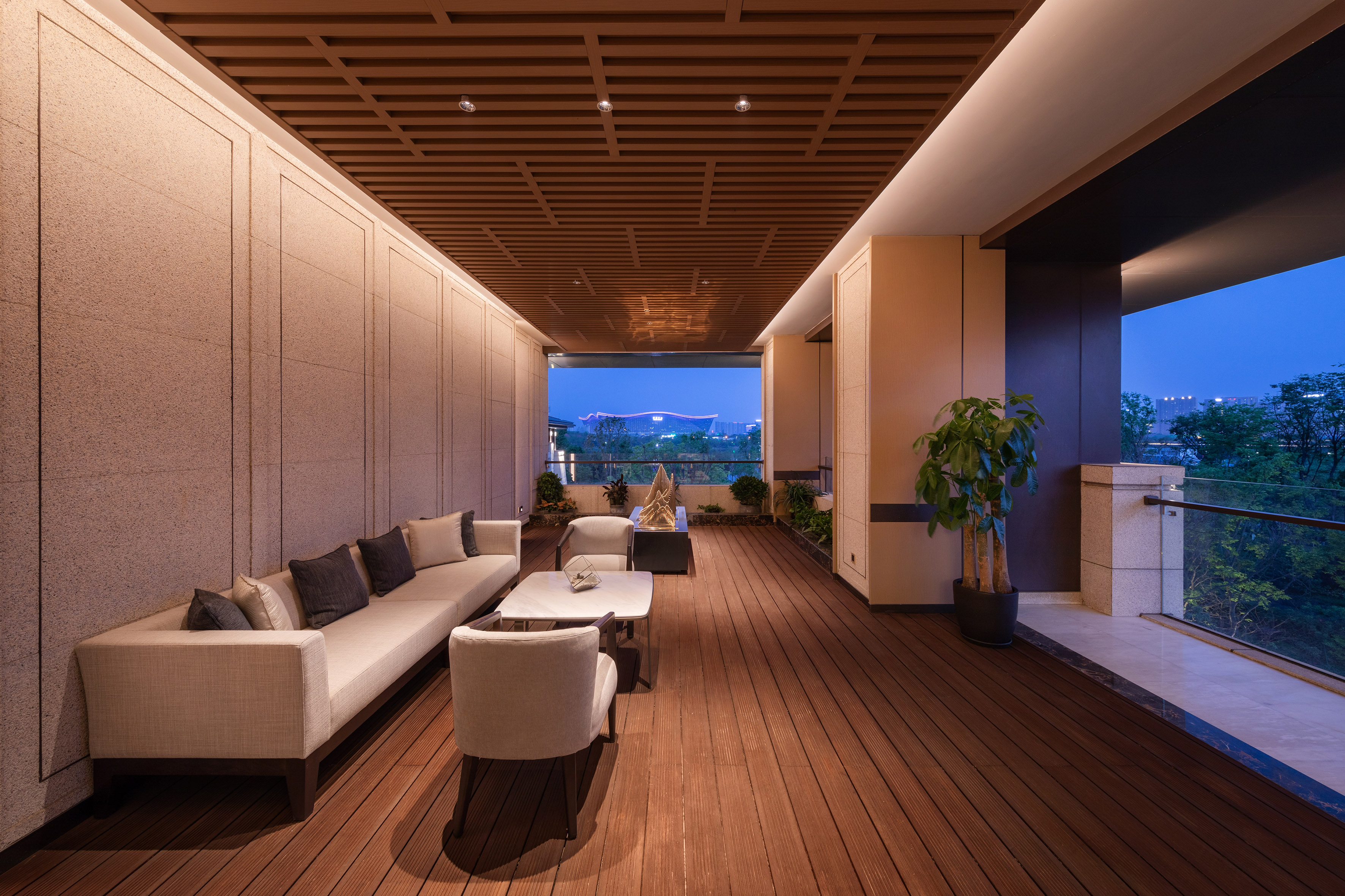 Modern outdoor lounge with sofa, wooden ceiling, and city view at dusk.