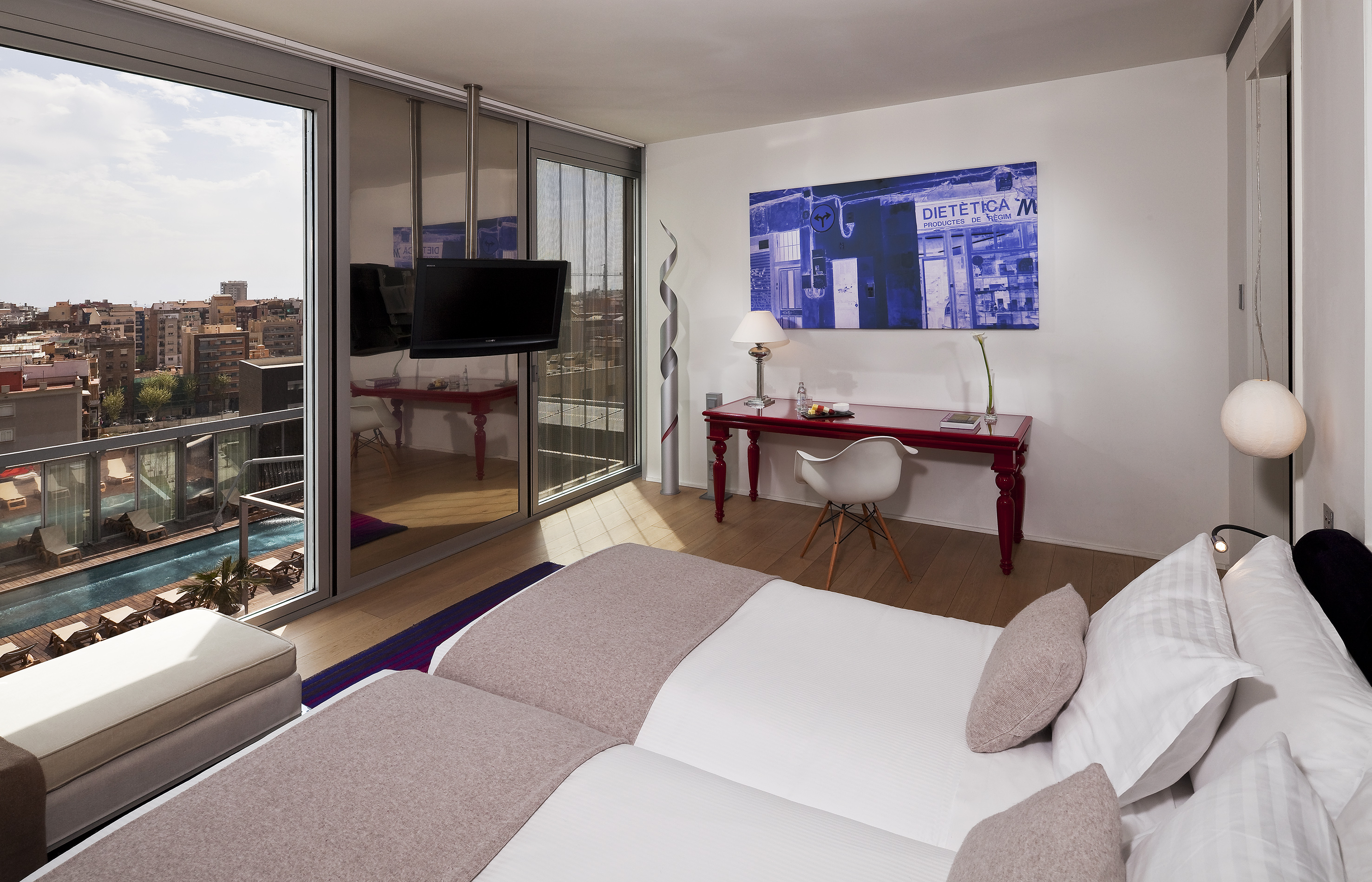Modern hotel room with city view, balcony pool, twin beds, and red desk.