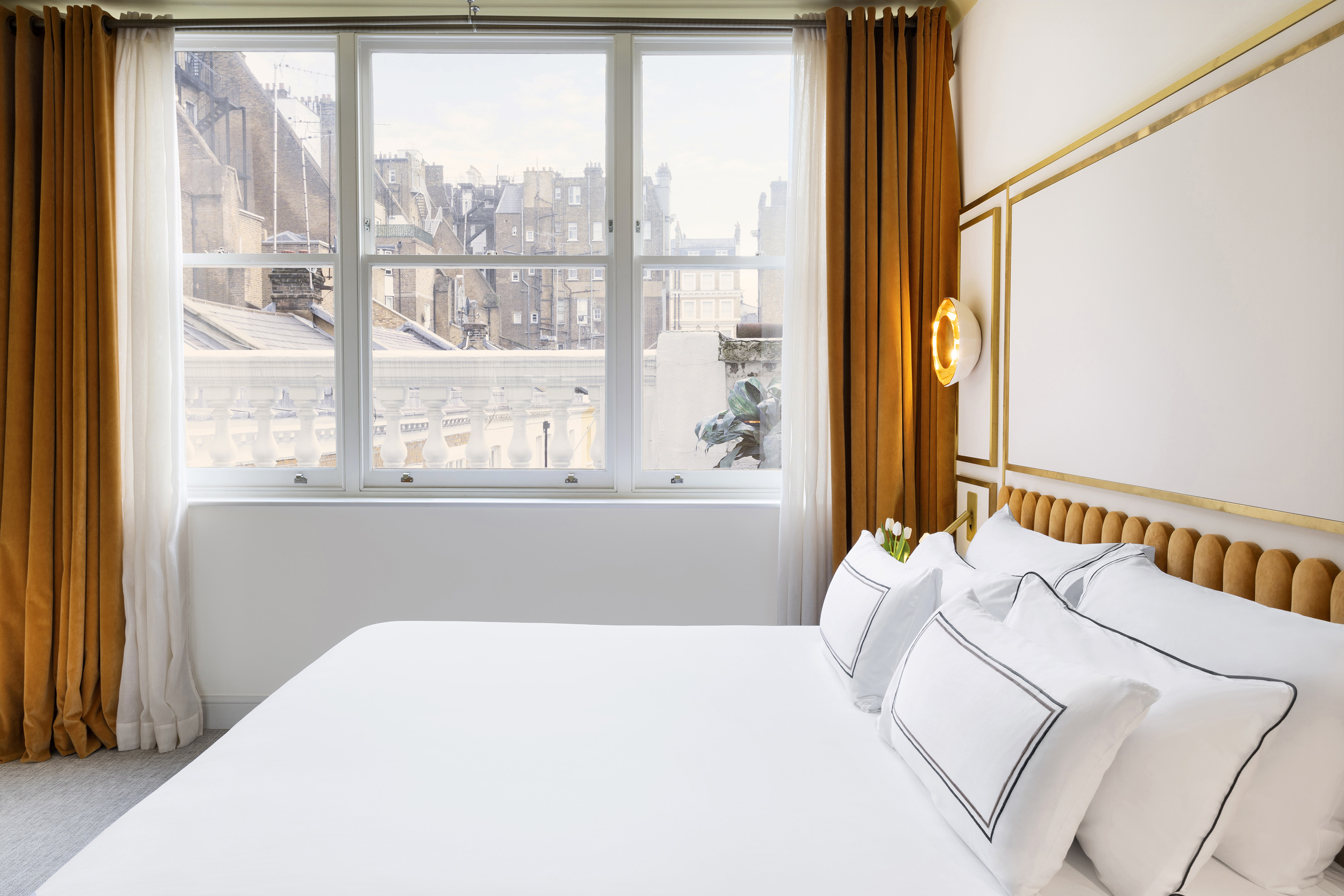 Modern hotel room with white bed, gold trim, velvet curtains, and city view.