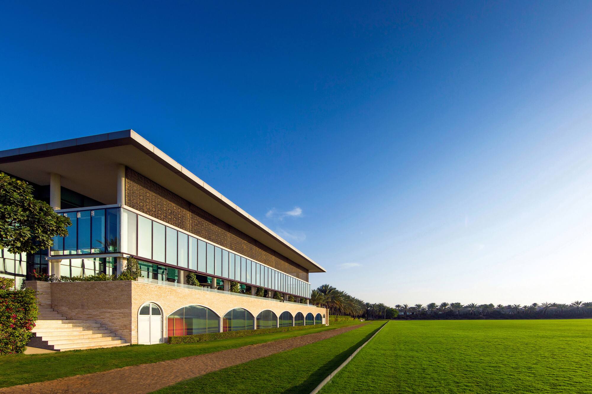 Modern building with glass facade overlooking a lush green field and palm trees.