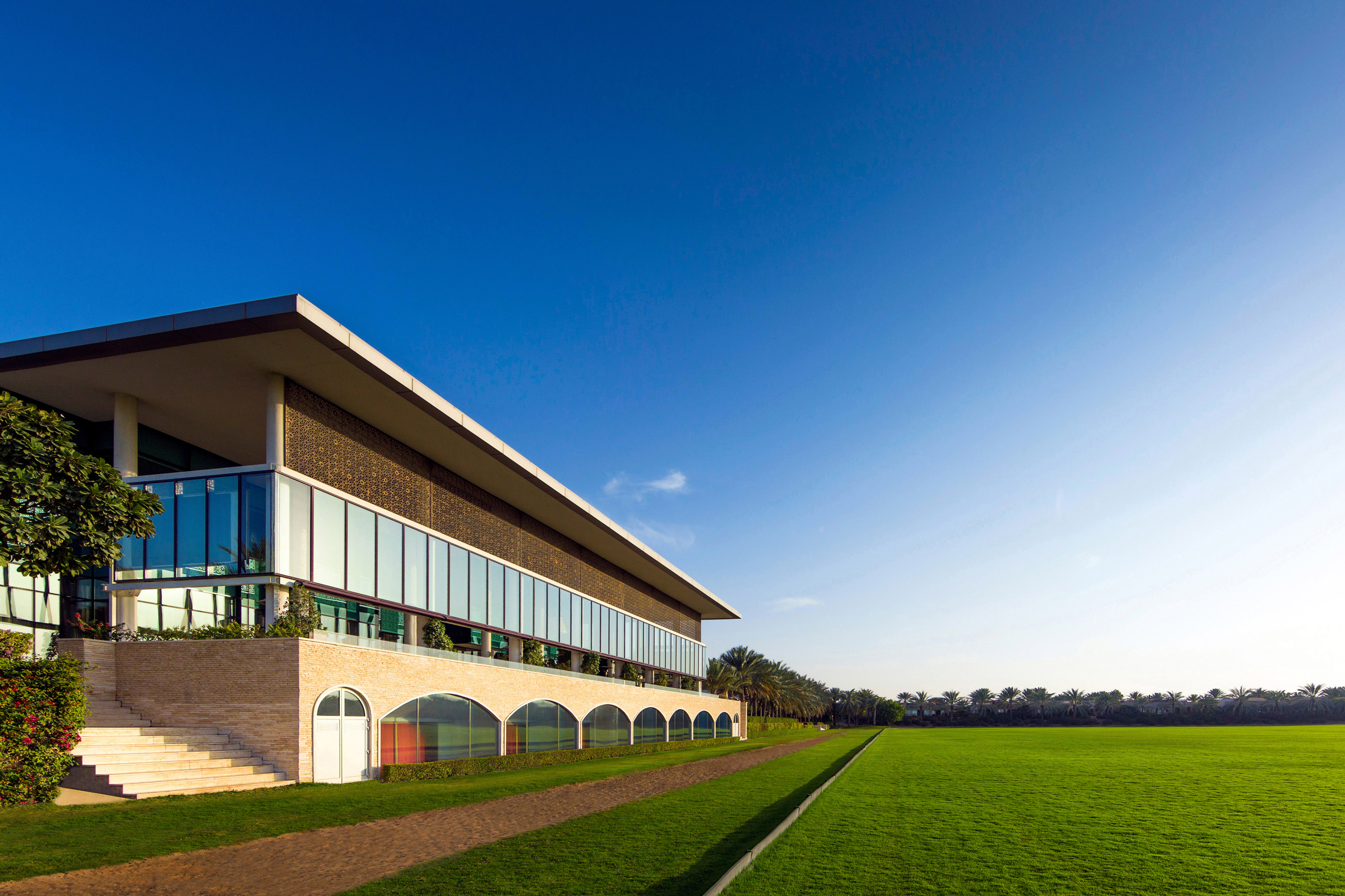 Modern building with glass facade overlooking a lush green field and palm trees.