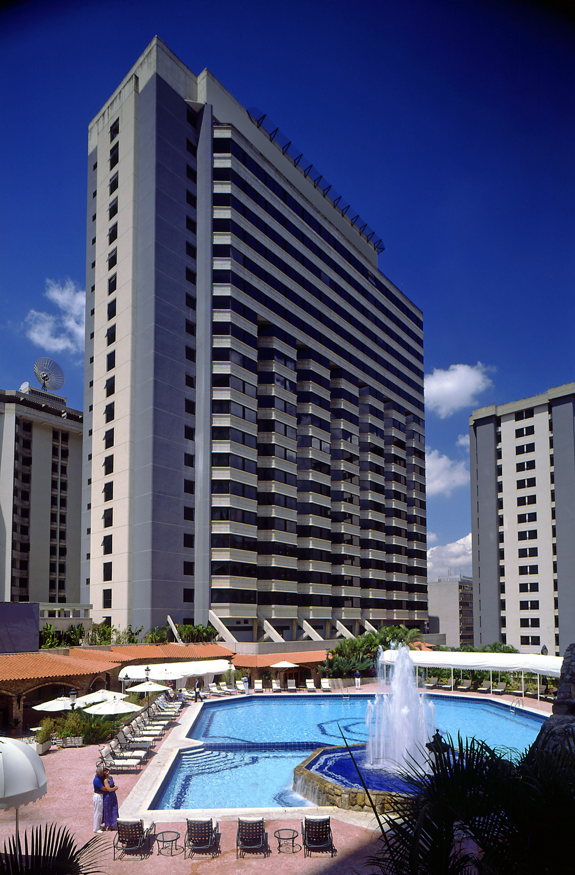 Modern high-rise hotel with outdoor resort pool, fountain, and sun loungers.