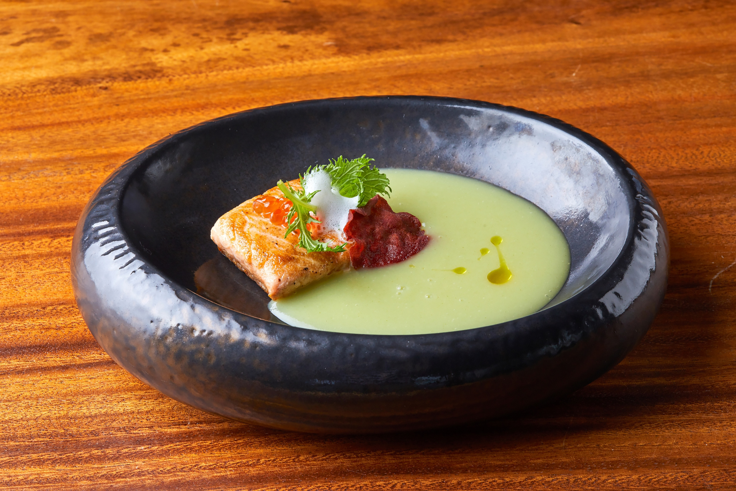 Seared salmon with green soup, red caviar, fresh herbs in a dark bowl.