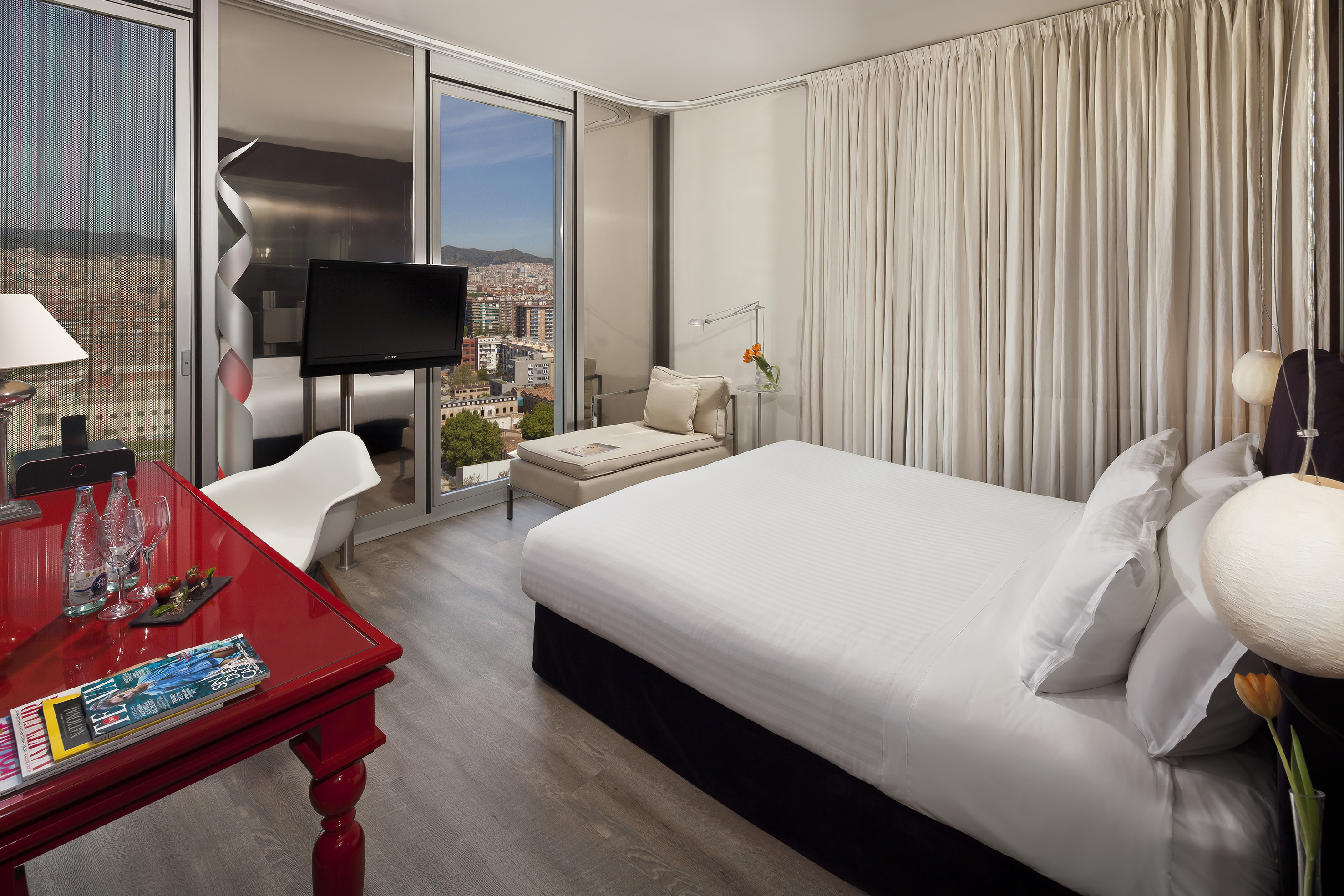 Modern hotel room with city view, large bed, red desk, and chaise lounge.