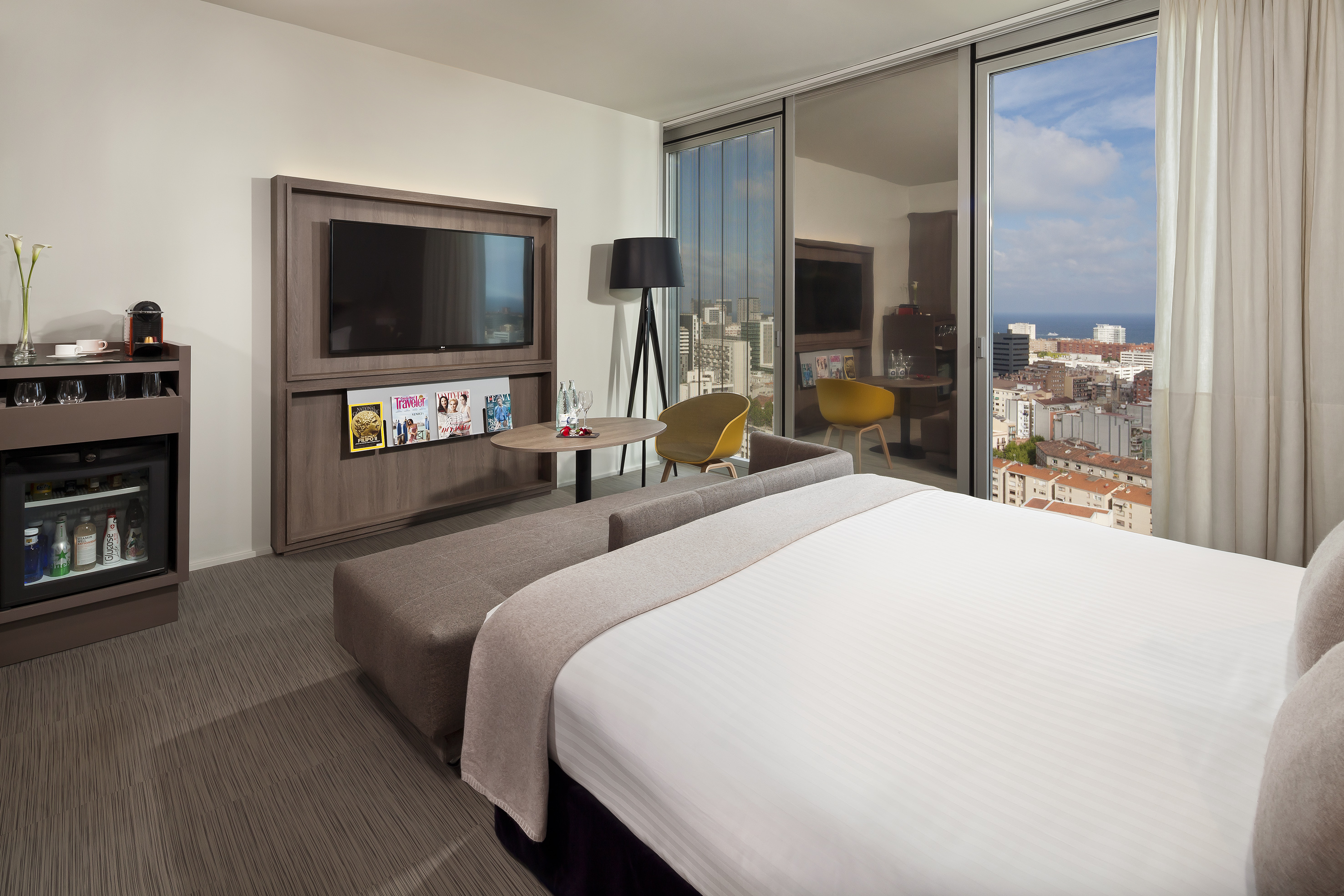 Modern hotel room with city and ocean view, bed, TV, minibar, and seating area.