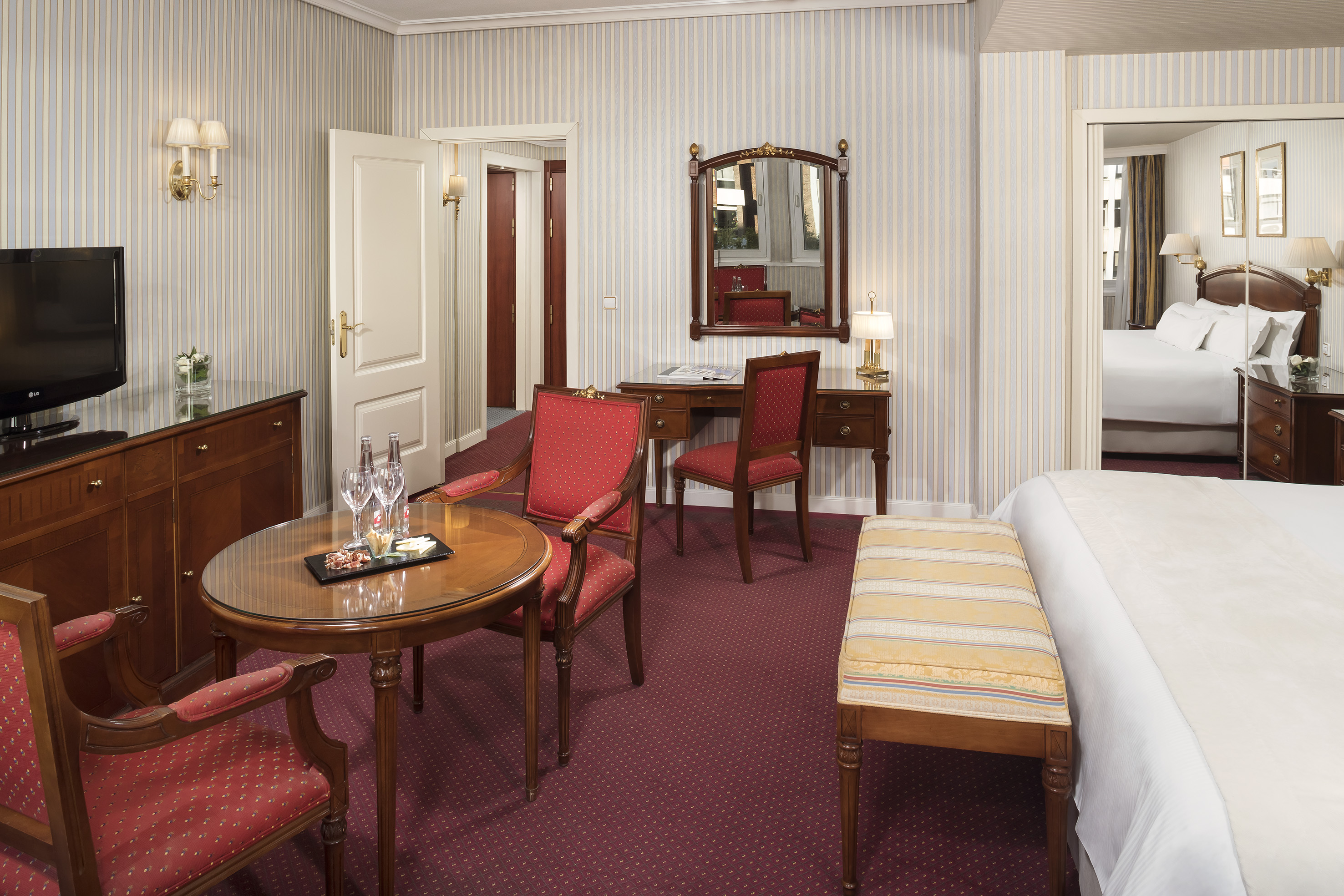 Classic hotel suite with red upholstered chairs, desk, and bedroom view.