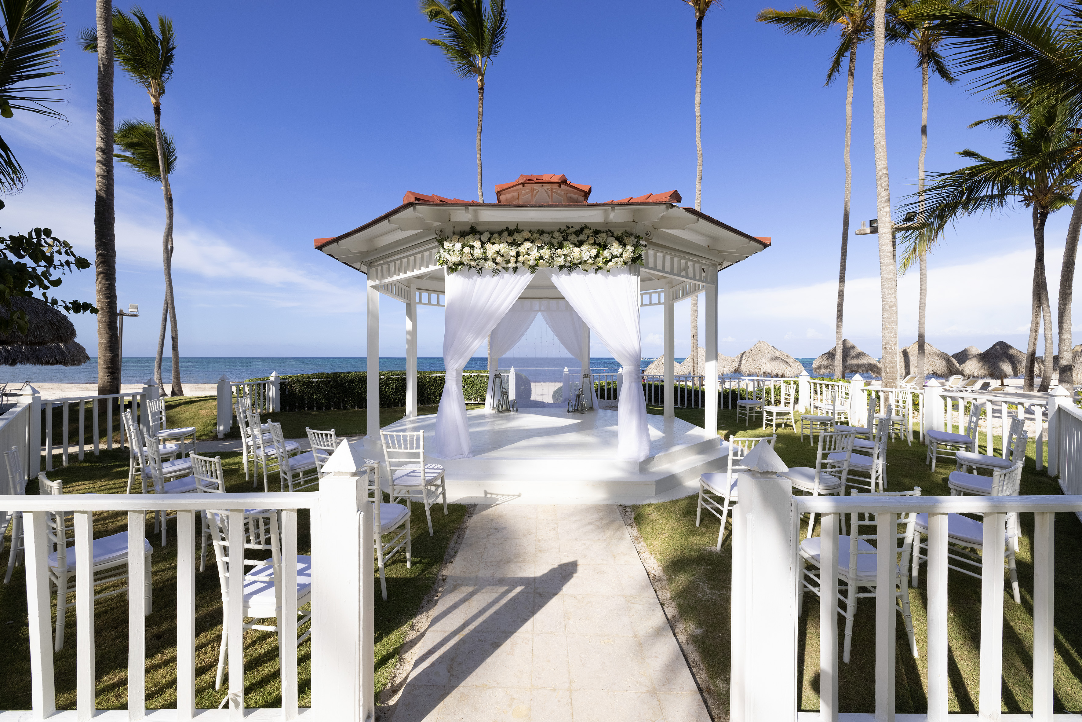 Beach wedding gazebo with white flowers, chairs, ocean, and palm trees.
