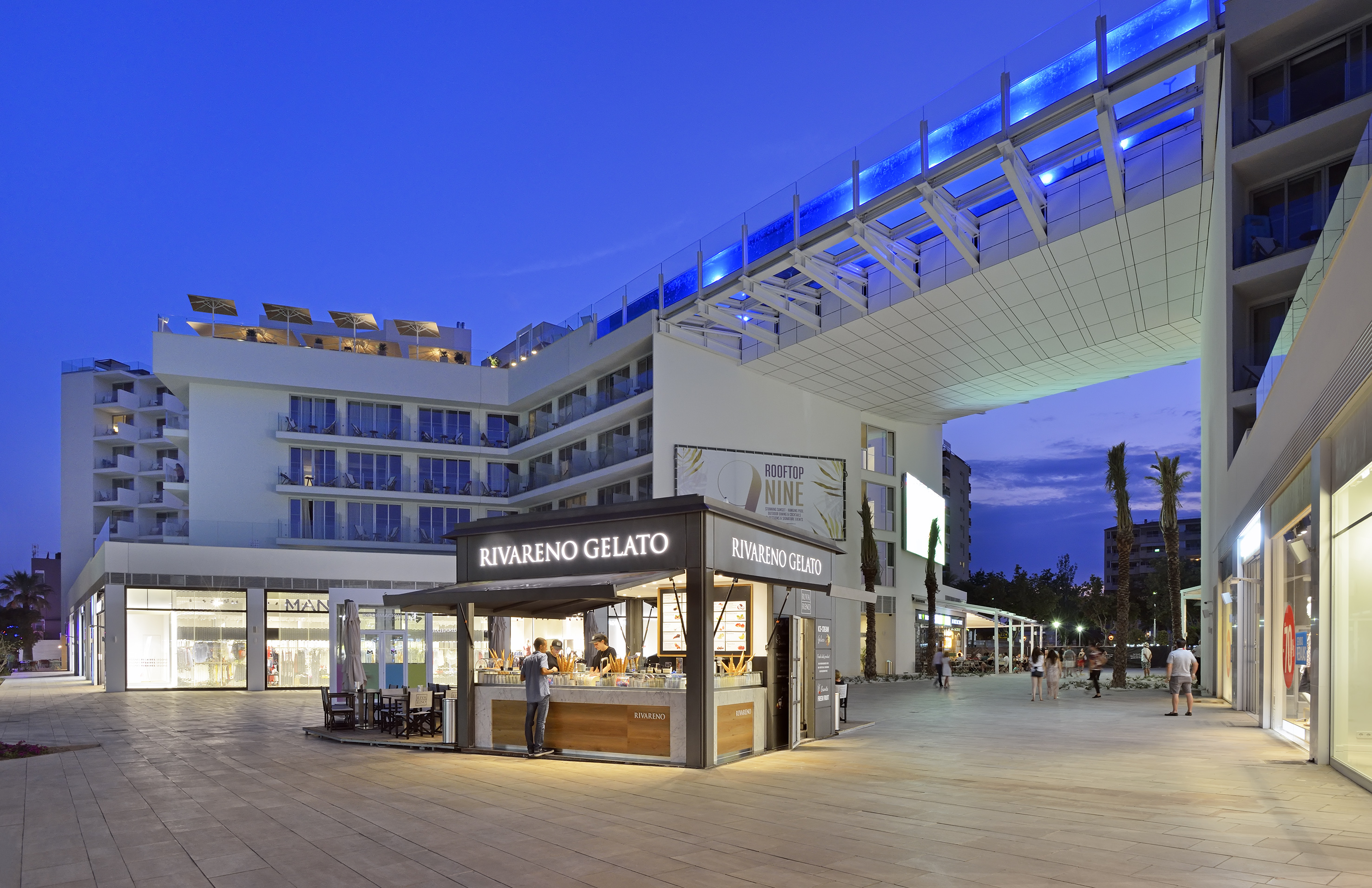 Rivareno Gelato shop in modern resort plaza with blue skybridge, evening.