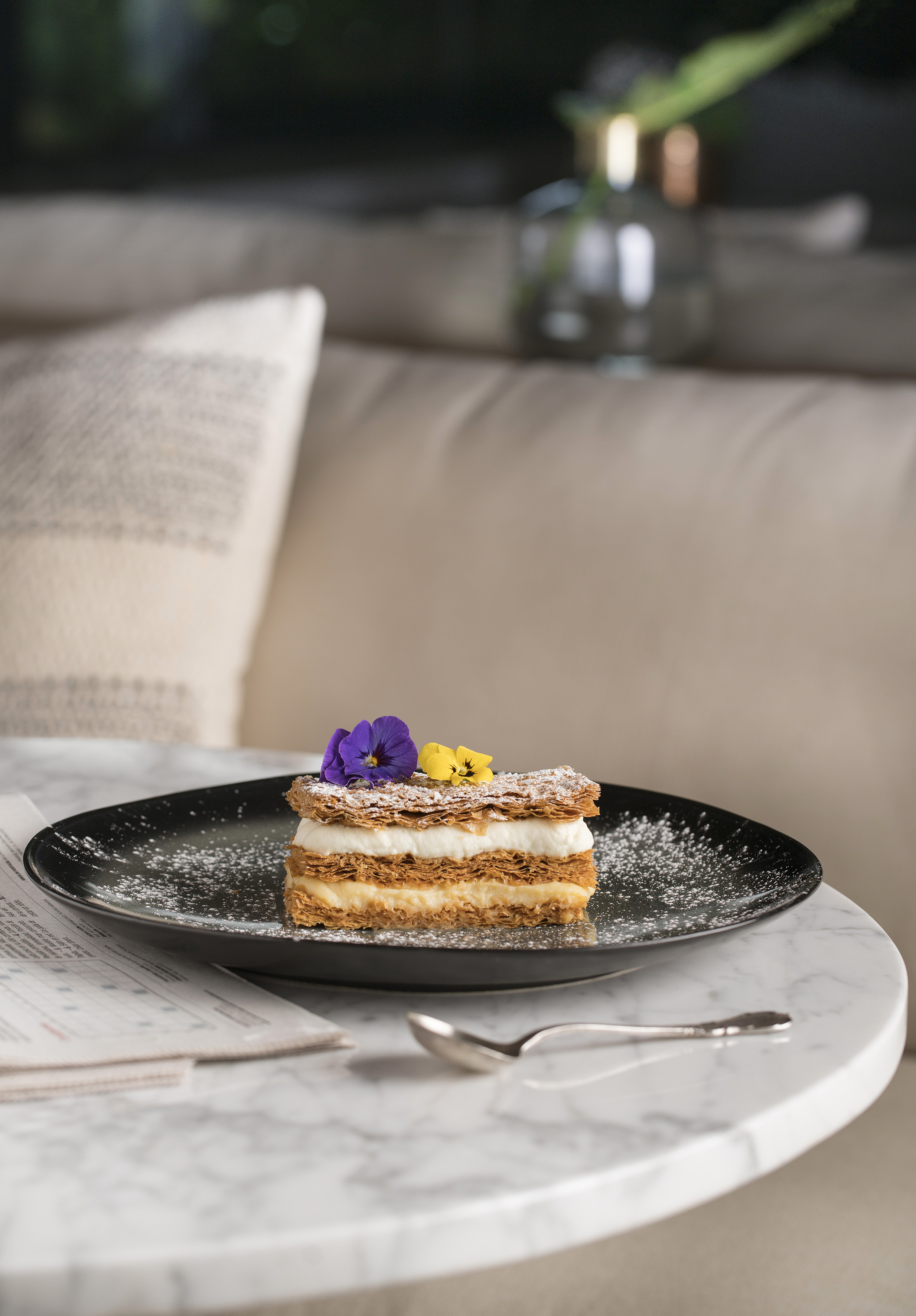 Mille-feuille with cream, powdered sugar, and edible flowers on black plate.