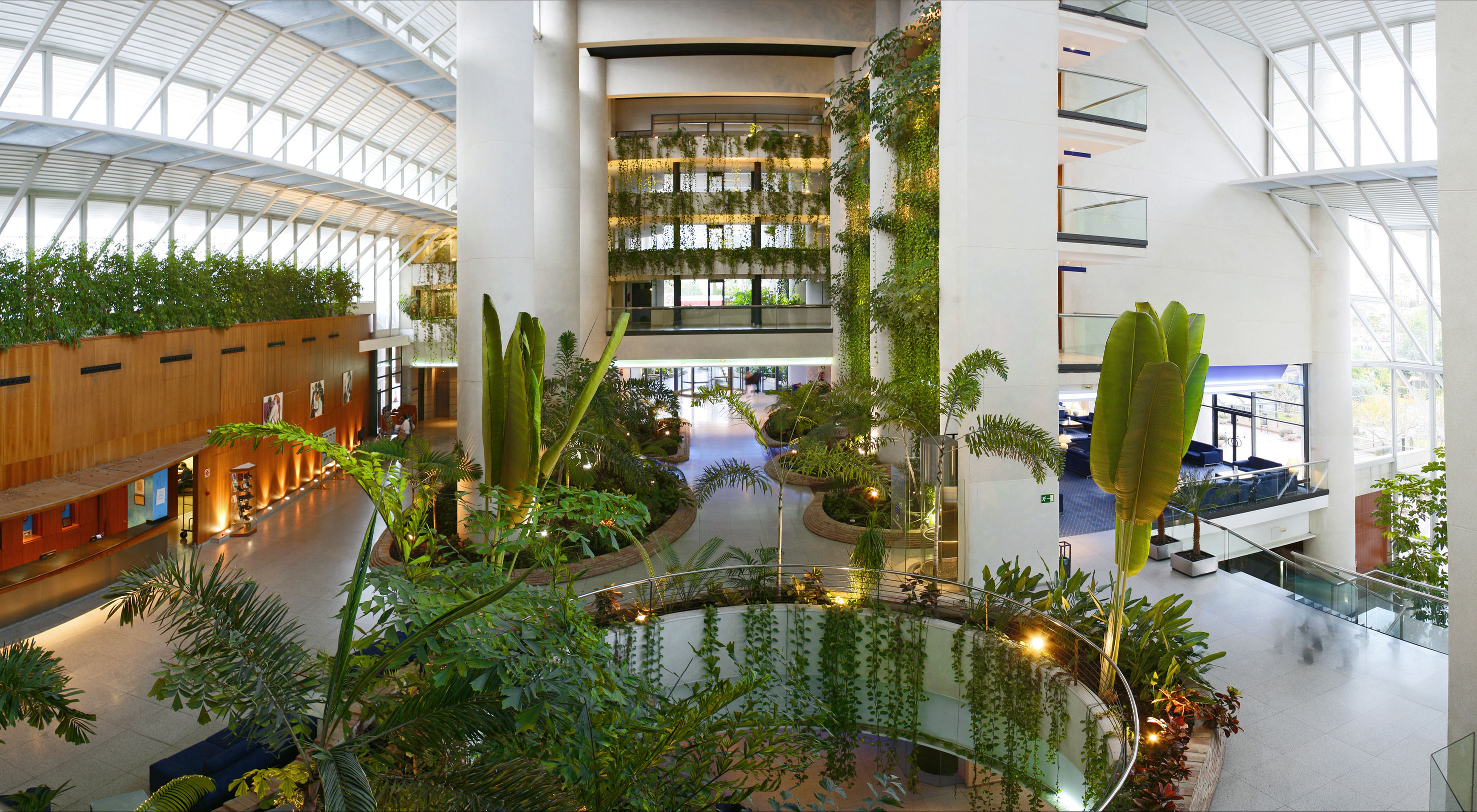 Modern hotel atrium with lush indoor garden, pond, and bright skylight.