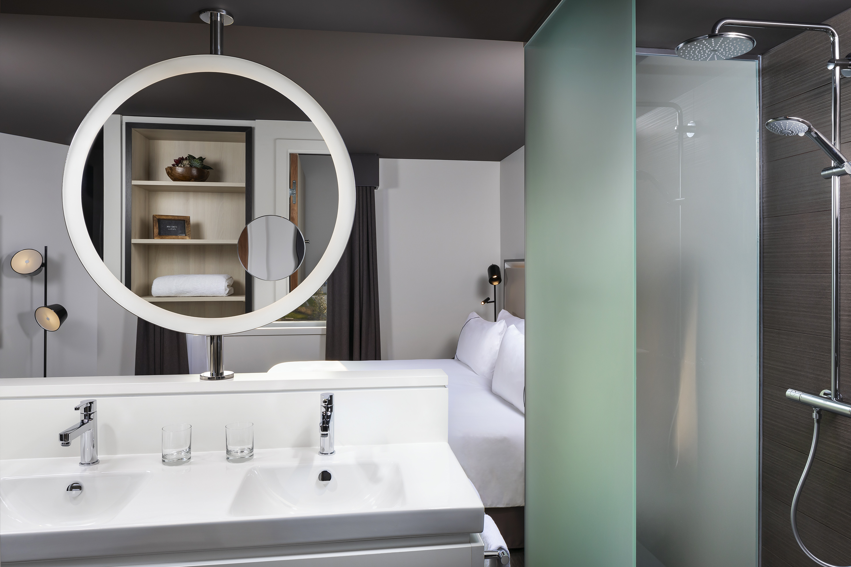 Modern hotel room interior with dual vanity, circular LED mirror, and glass shower.