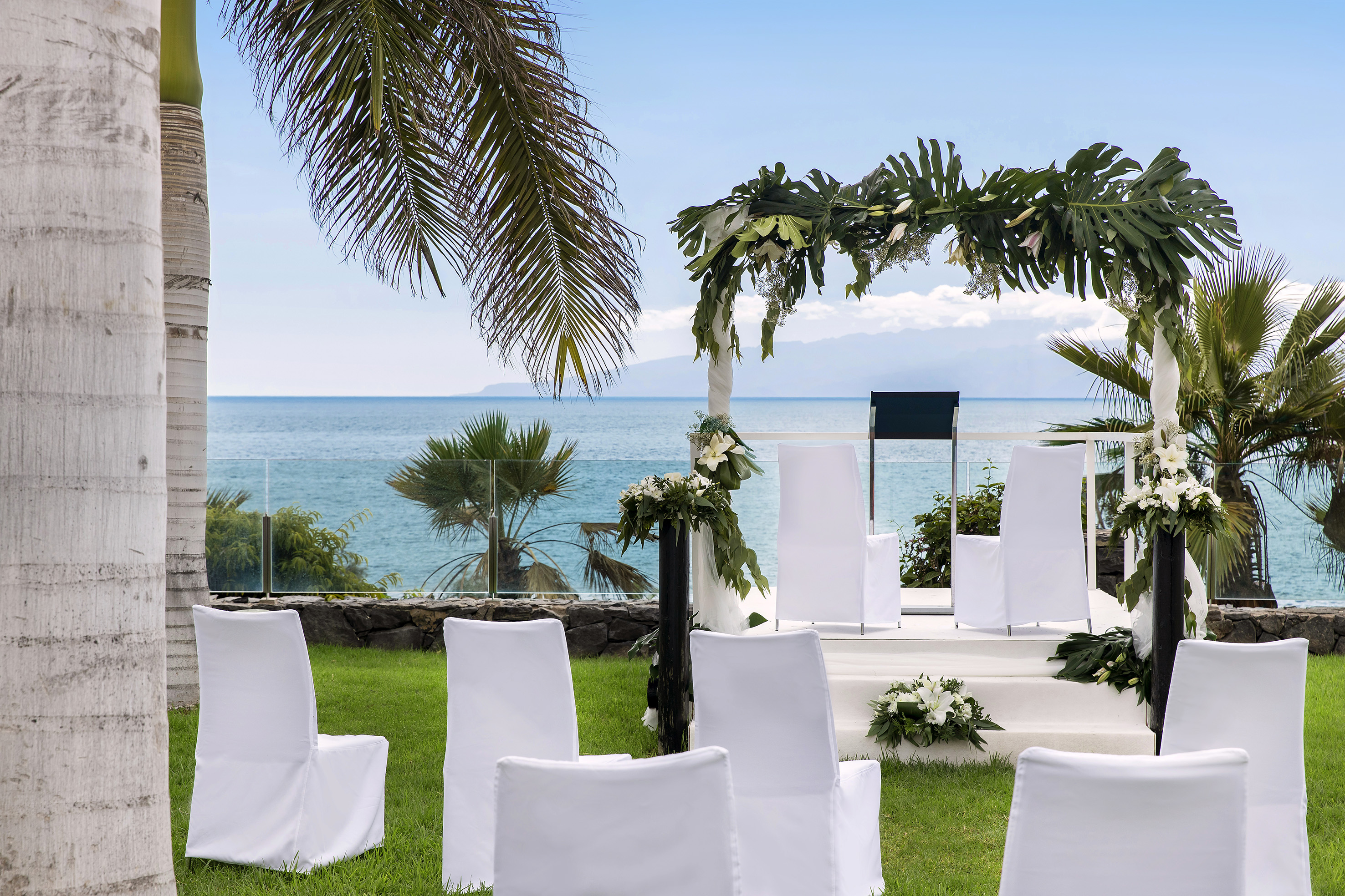 Tropical ocean view wedding ceremony with white chairs and floral arch.