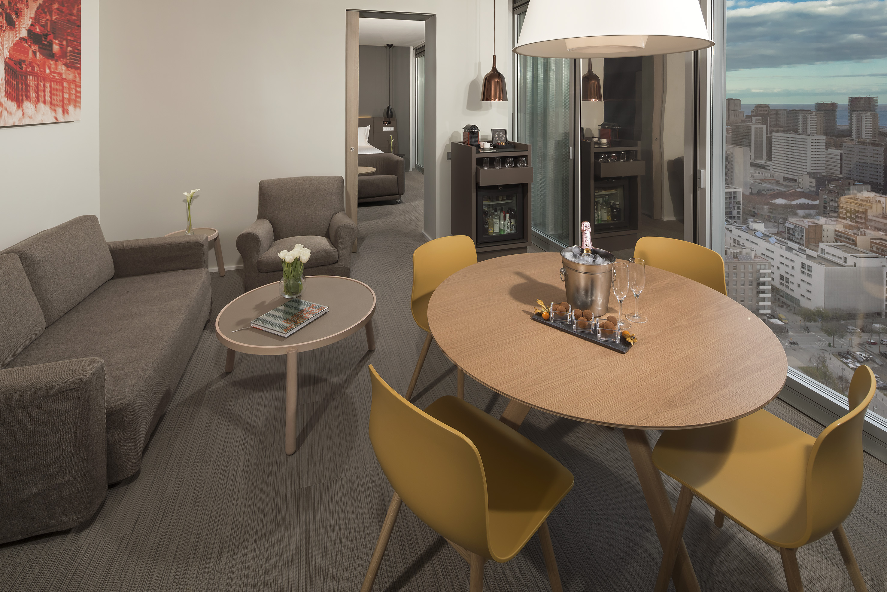 Contemporary hotel suite with city view, living area, dining table with champagne.