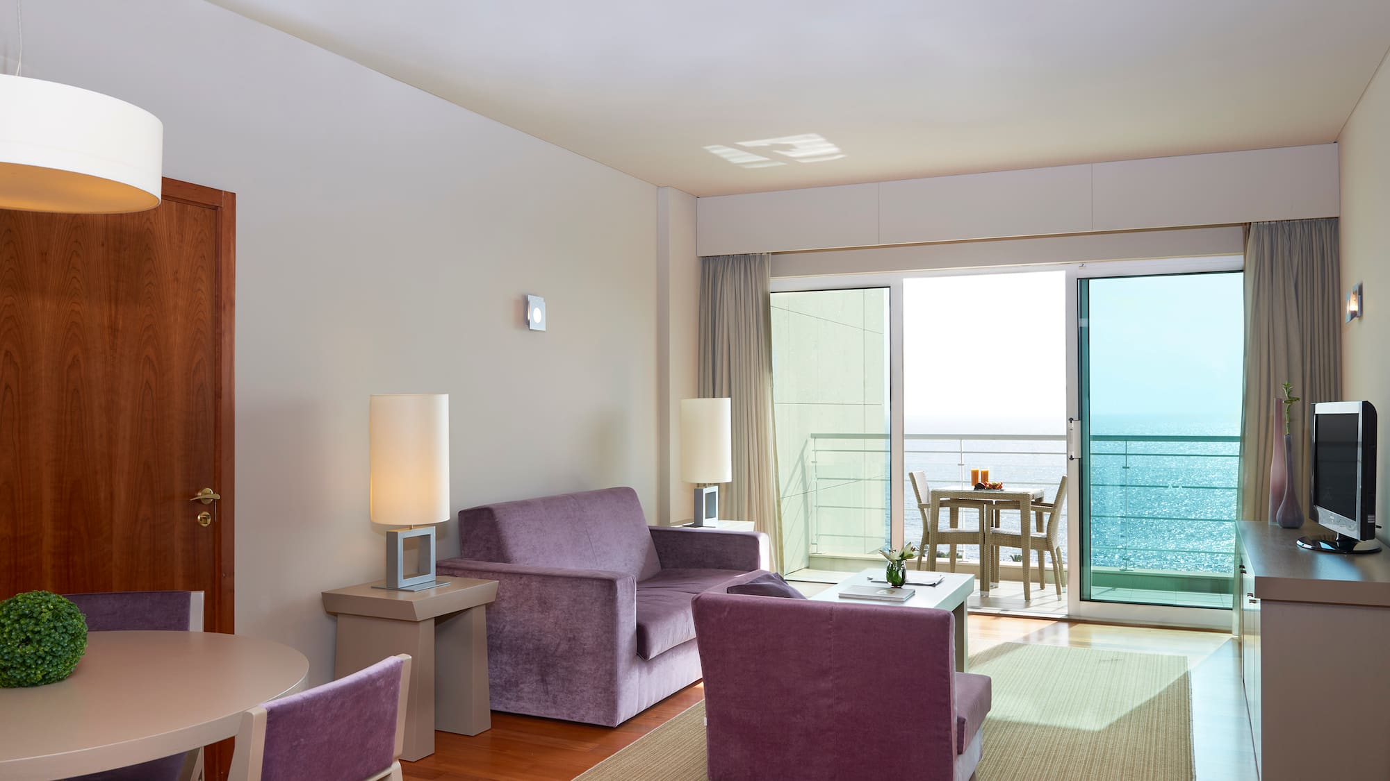 Modern hotel living room with purple furniture, dining area, and ocean view balcony.