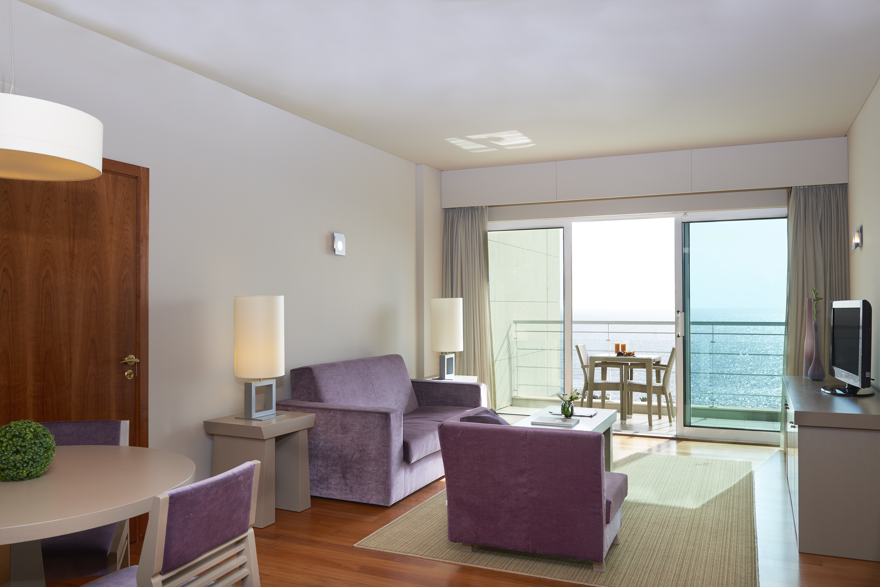Modern hotel living room with purple furniture, dining area, and ocean view balcony.