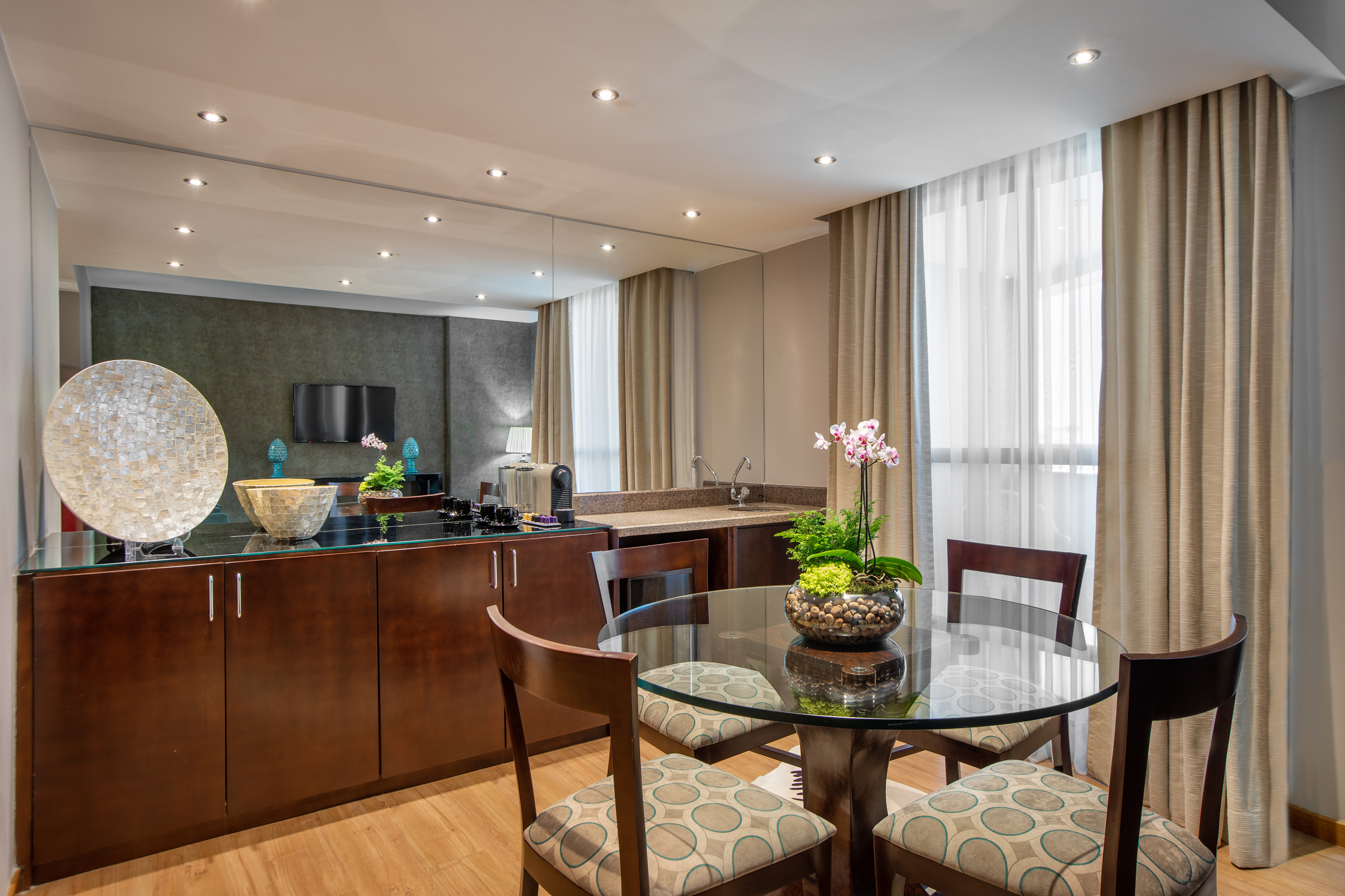 Modern hotel suite dining area, glass table, chairs, kitchenette.