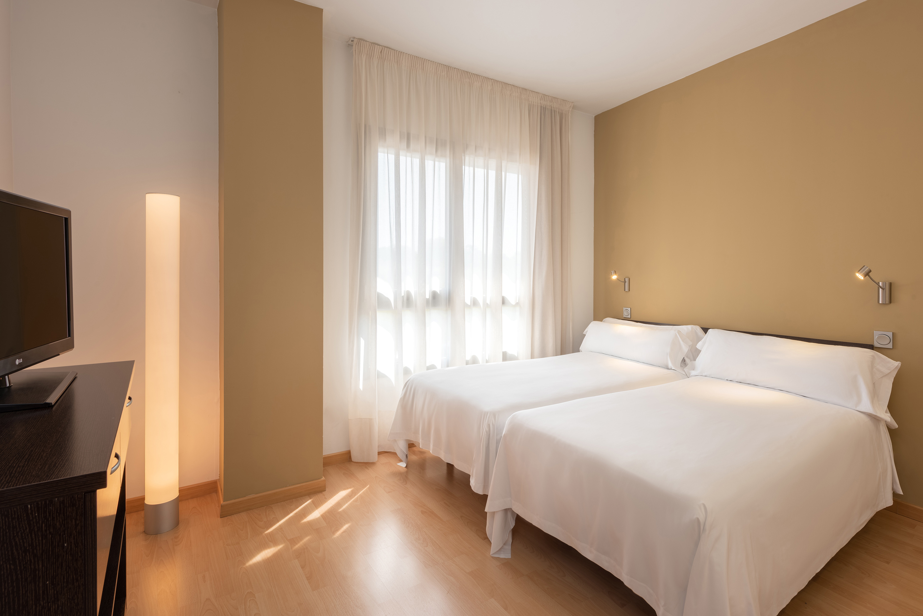 Modern hotel guest room with two twin beds, wood floor, bright window, and TV.