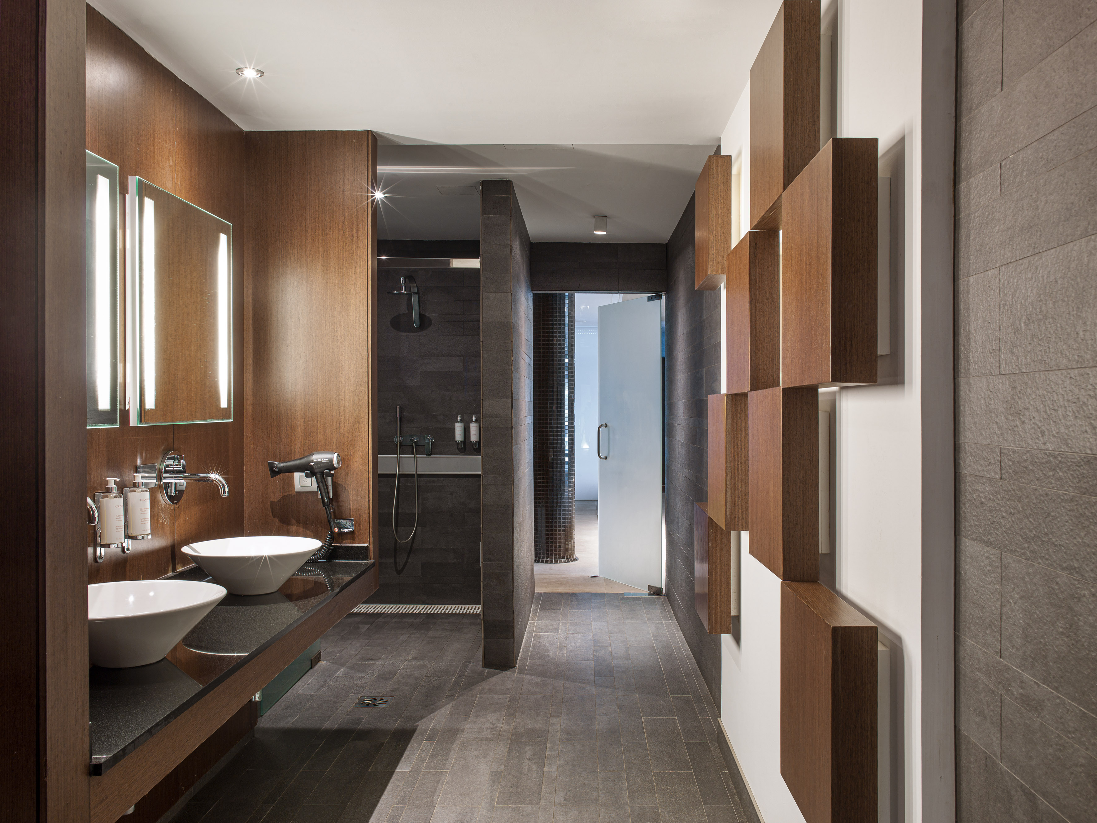 Modern spa bathroom with twin sinks, wood vanity, and dark tiled shower area.