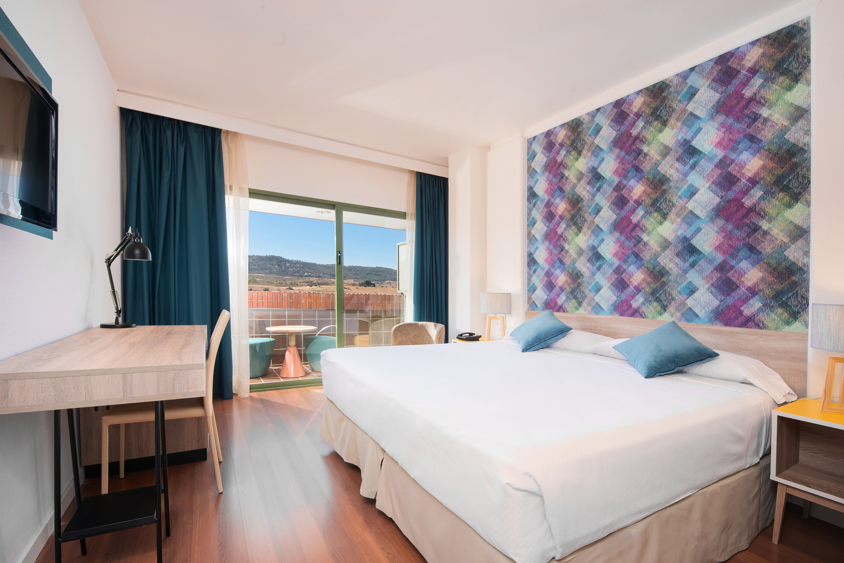 Hotel room with king bed, geometric wall, desk, and balcony mountain view.