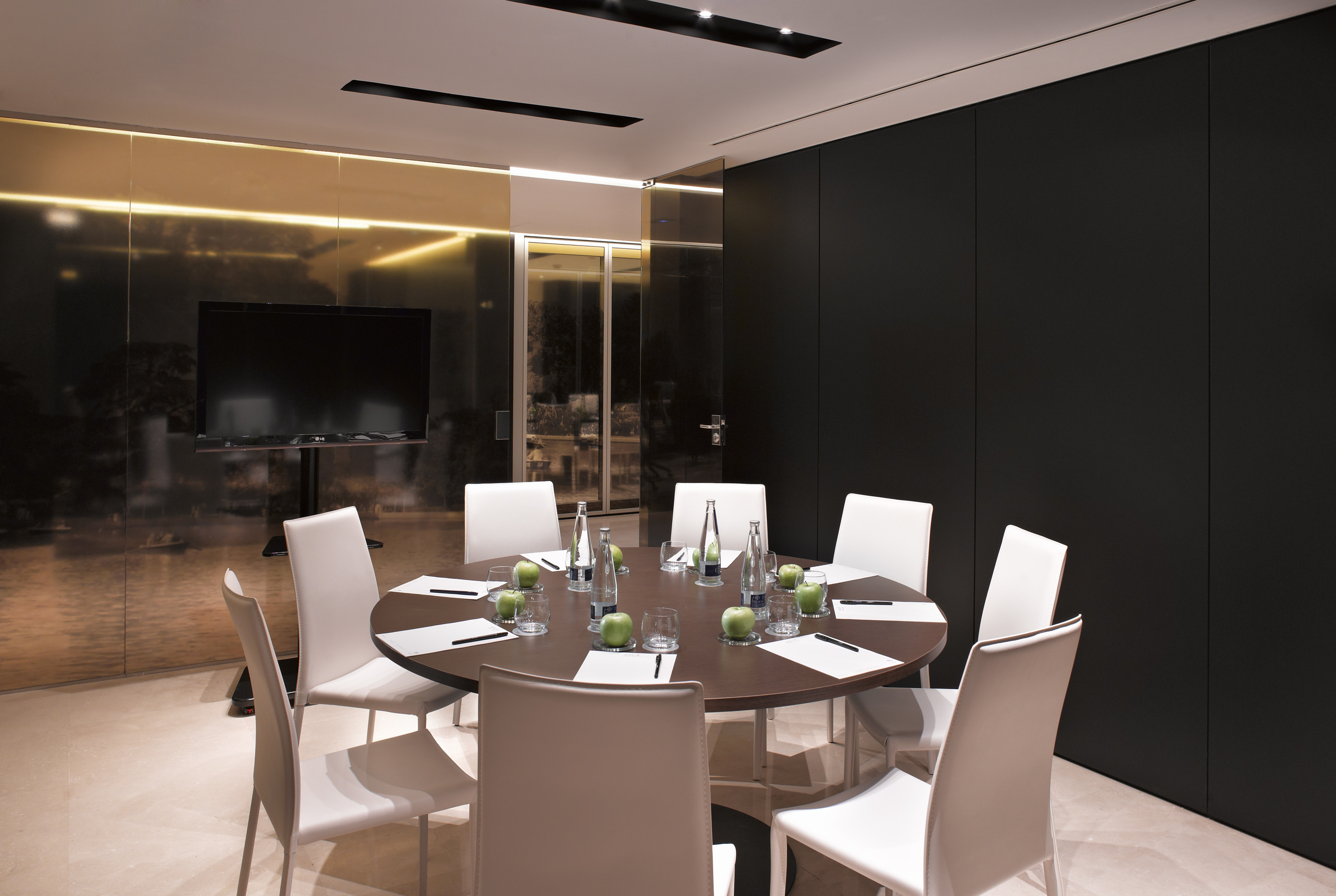 Modern boardroom with round table, white chairs, water bottles, and TV.