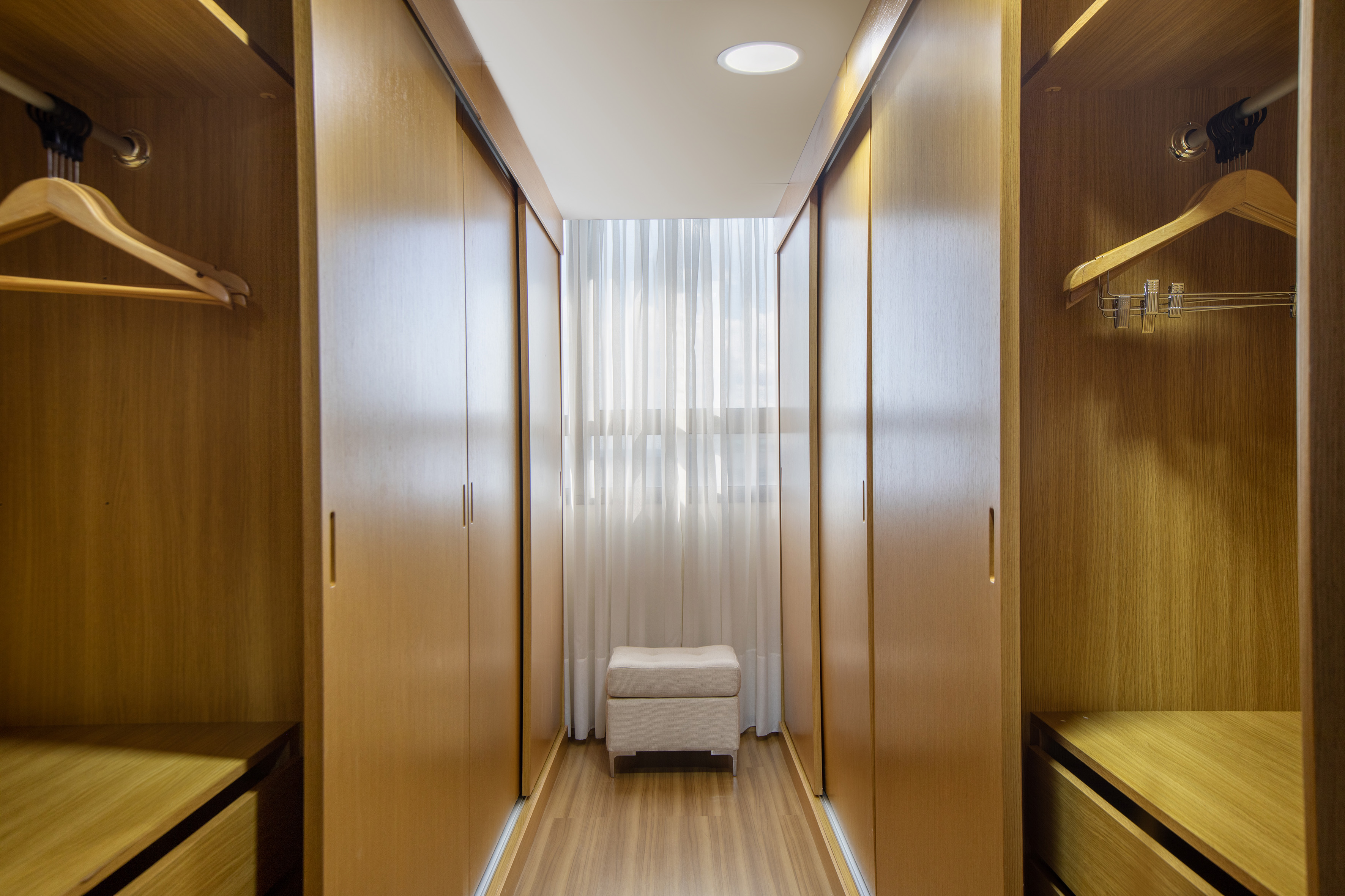 Modern walk-in closet with light wooden built-in cabinets and central ottoman.