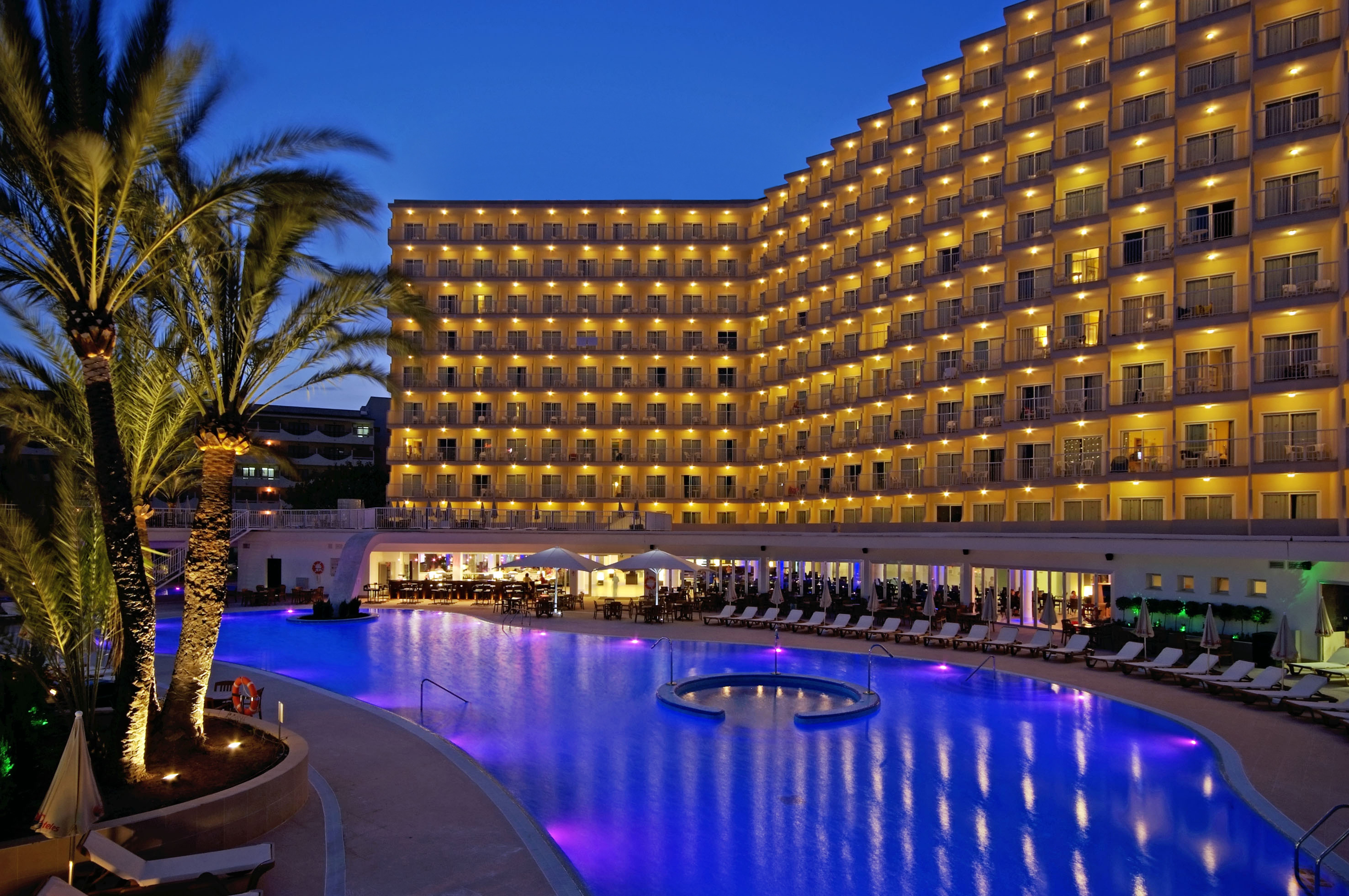 Hotel resort with illuminated balconies, glowing blue pool, and palm trees at night.